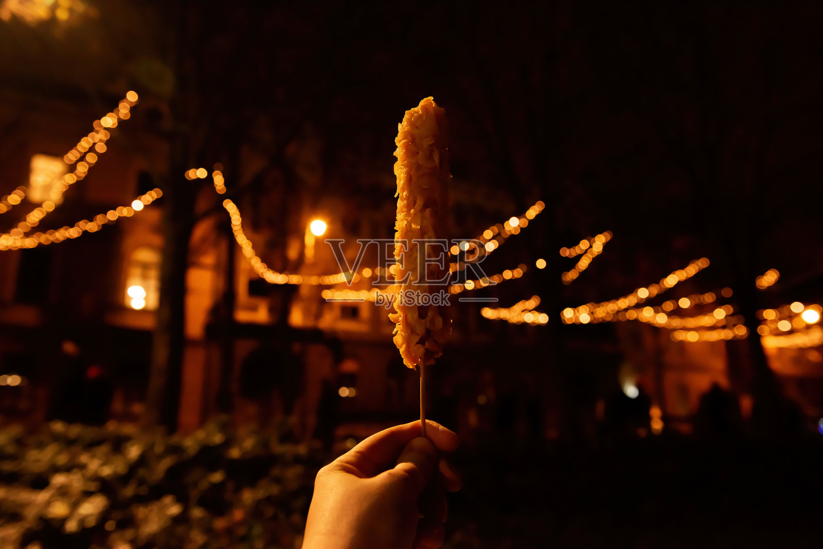 在城市圣诞装饰的背景下，手拿一根木棒，一只玉米热狗片。照片摄影图片