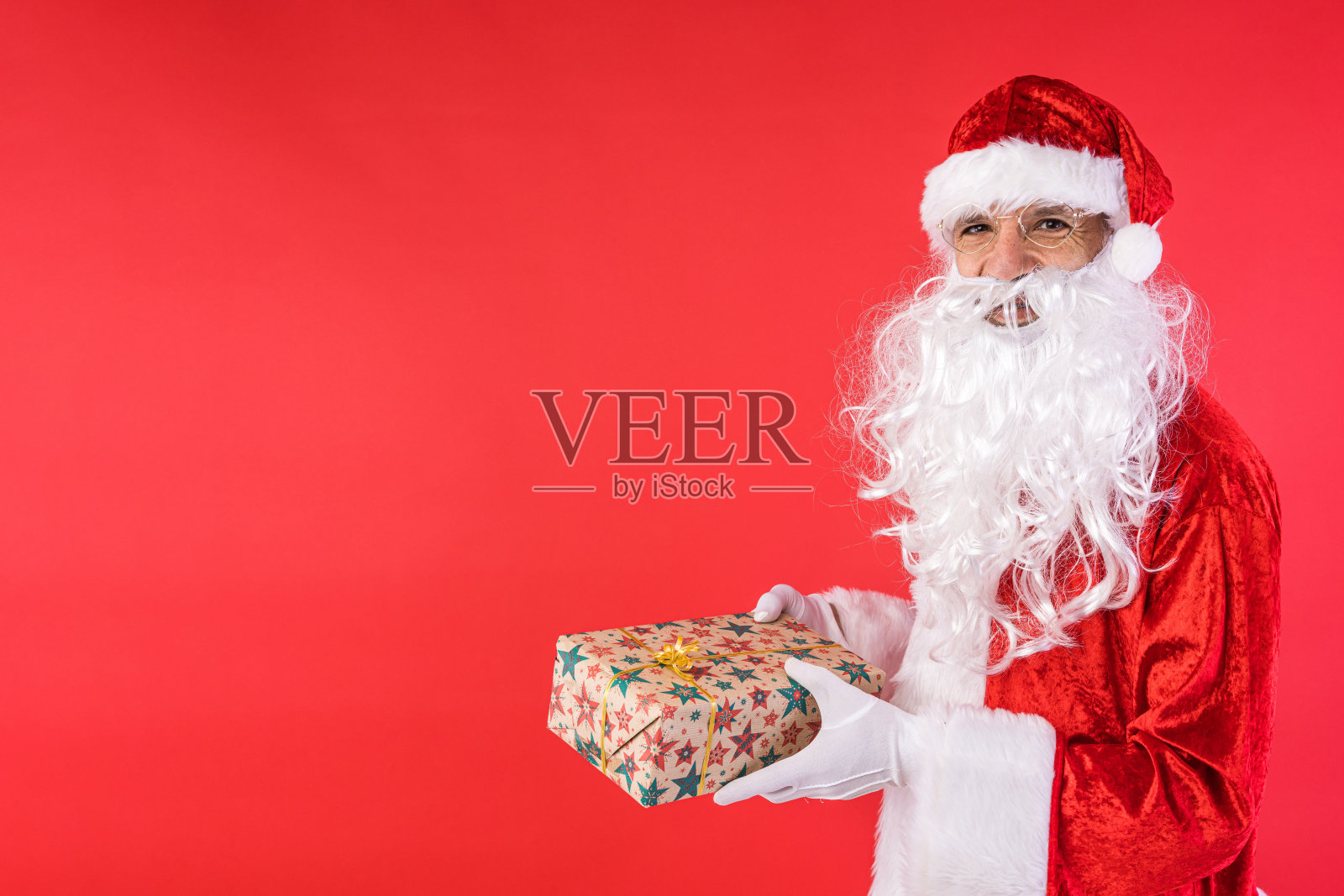 一名装扮成圣诞老人的男子拿着一份礼物，旁边有复印空间，红色背景。圣诞节，庆祝，礼物，消费主义和幸福的概念。照片摄影图片