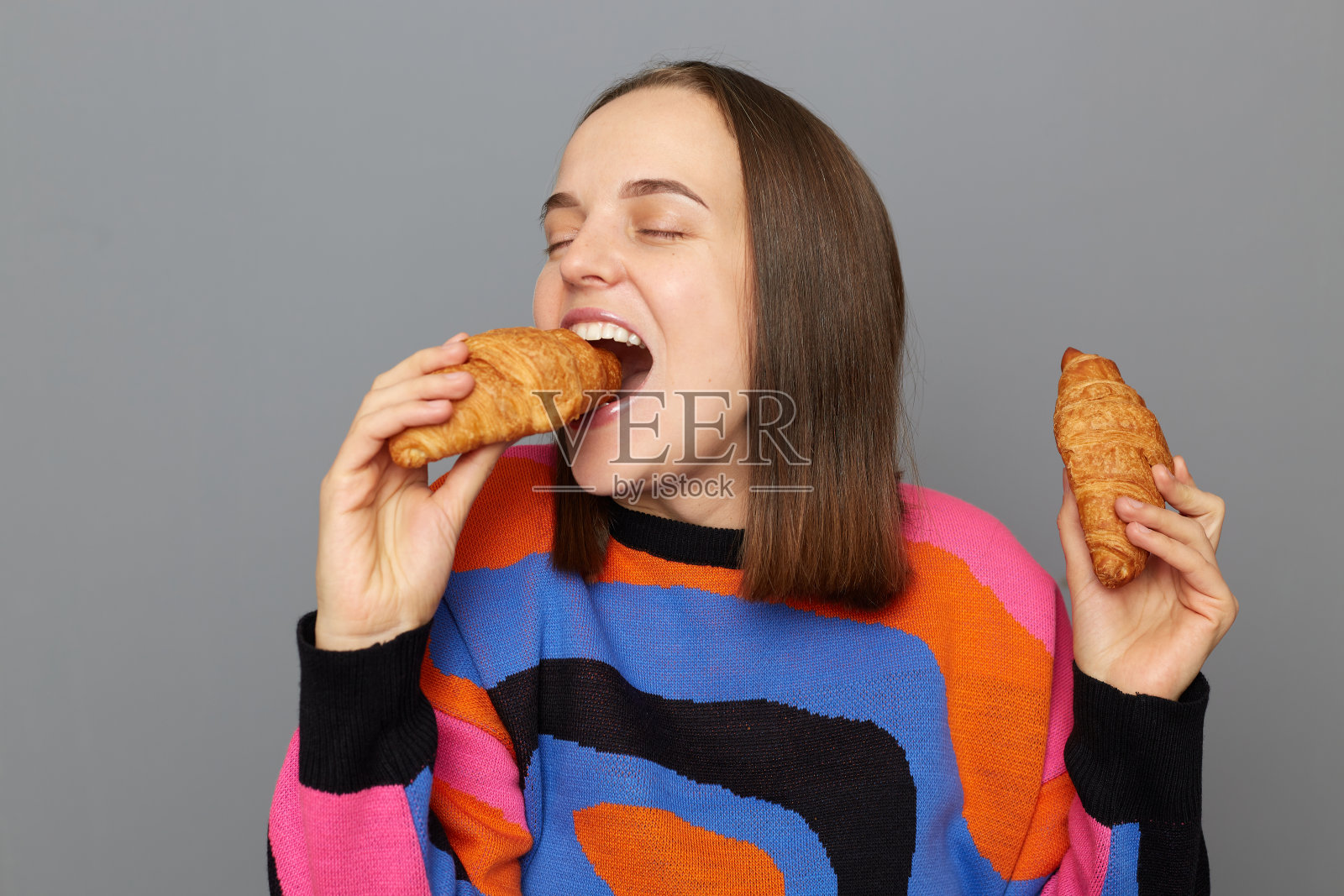 室内拍摄的快乐正面白人女性穿着毛衣，在灰色背景下摆出孤立的姿势，咬着羊角包，感觉饿了，喜欢吃甜食。照片摄影图片