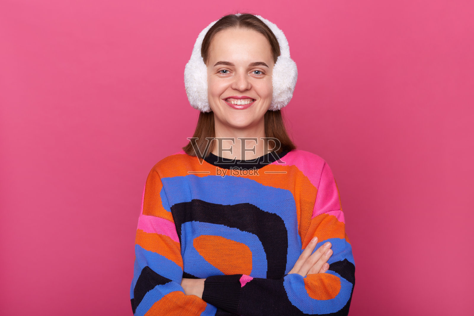 室内拍摄的微笑满意的年轻成年女子戴着蓬松的冬季耳套和温暖的毛衣站在粉红色的背景，交叉双臂孤立，看着相机与快乐的表情。照片摄影图片