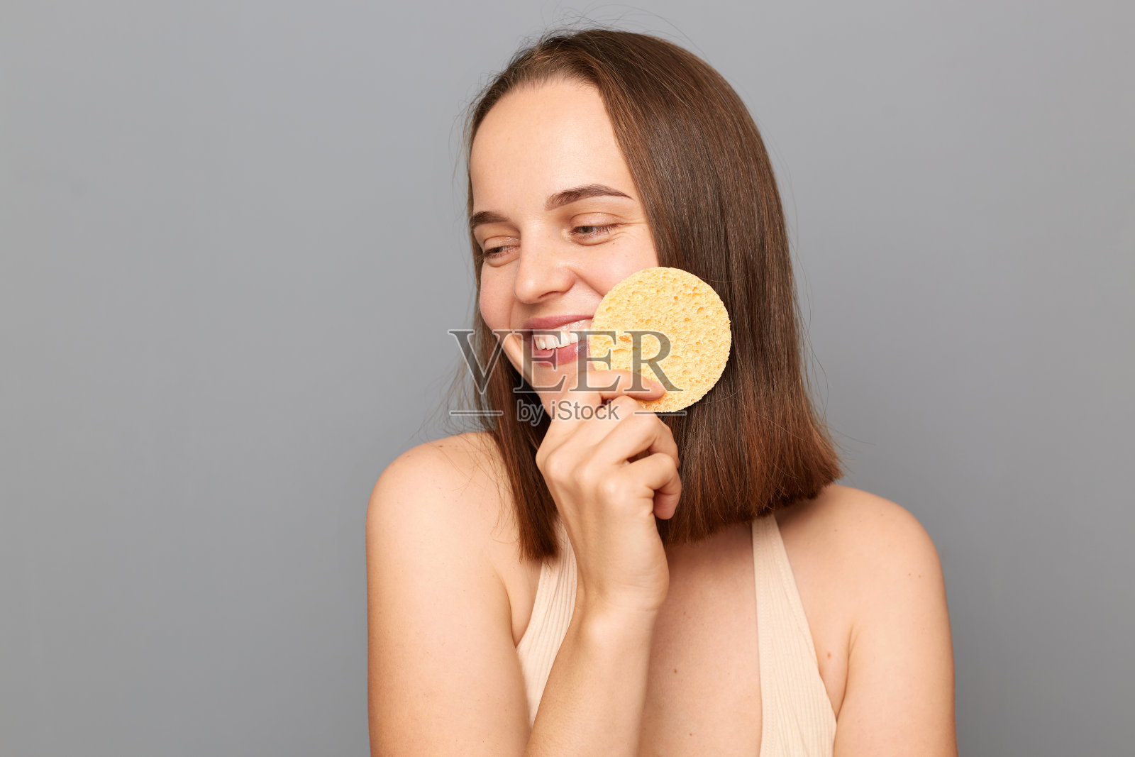 快乐的年轻女孩的照片，温柔的微笑抱着化妆海绵在灰色背景上孤立的脸，看向别处，享受美容手术。照片摄影图片