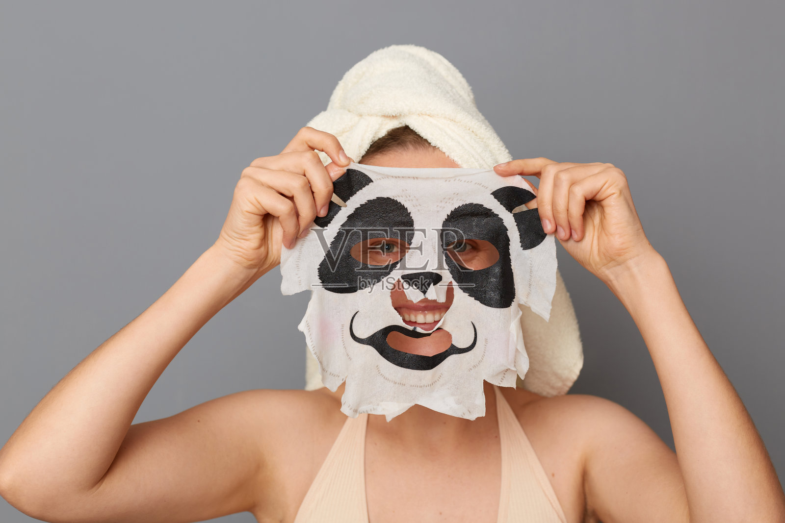 迷人的年轻女子在室内拍摄，头上裹着毛巾，敷着熊猫嘴形的面膜，做美容手术。照片摄影图片