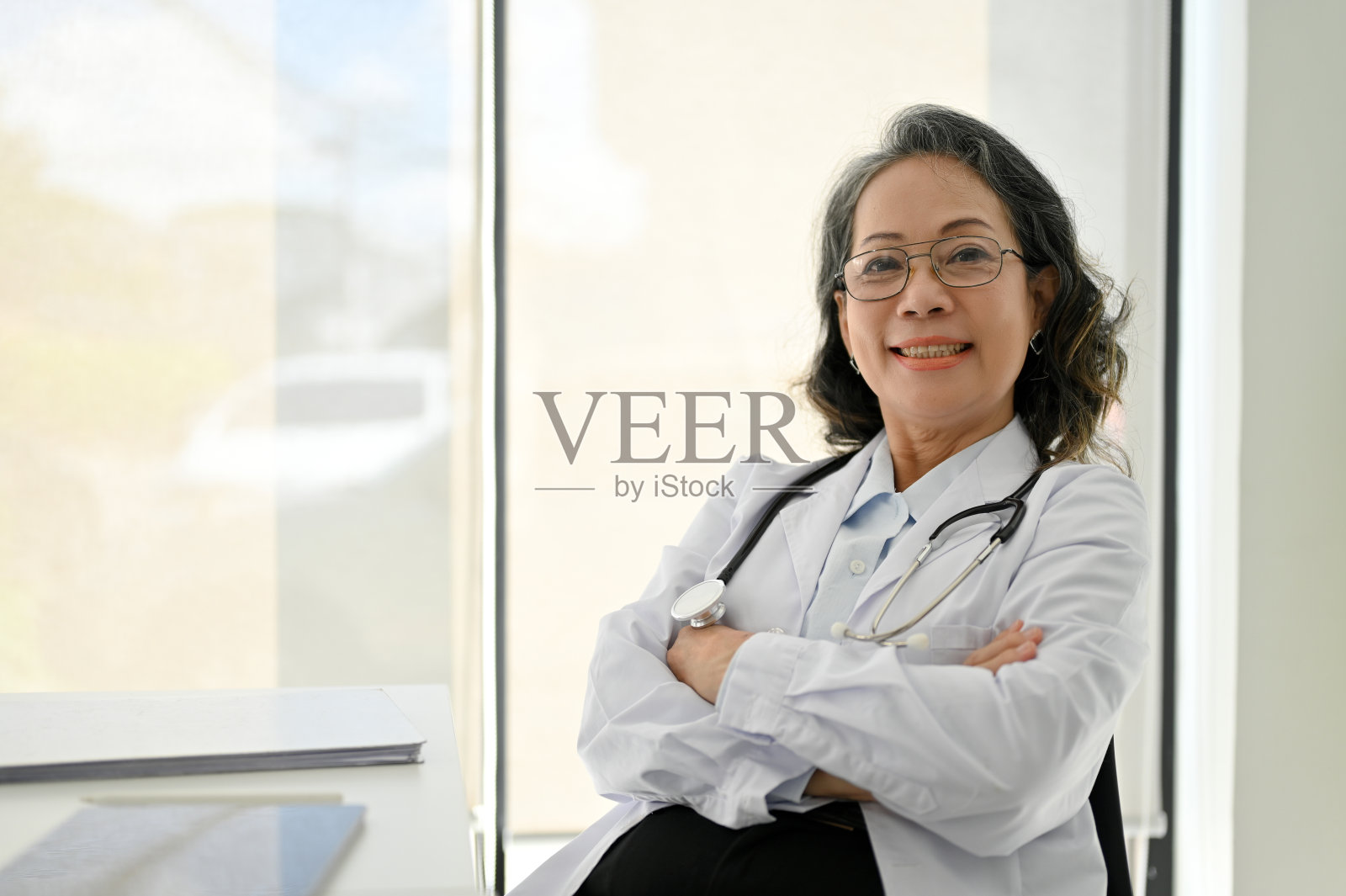 成功的亚裔女医生坐在窗边，双手交叉，看着镜头。照片摄影图片