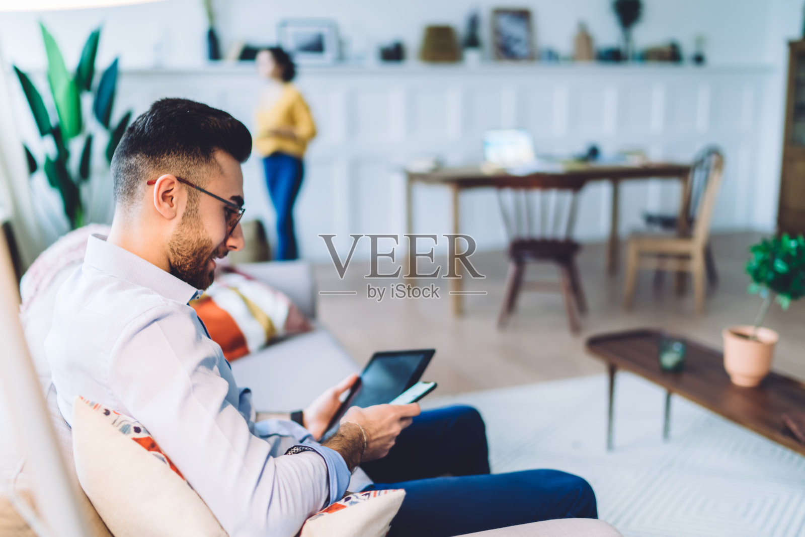 年轻的少数民族男子坐在沙发上使用平板电脑和智能手机照片摄影图片