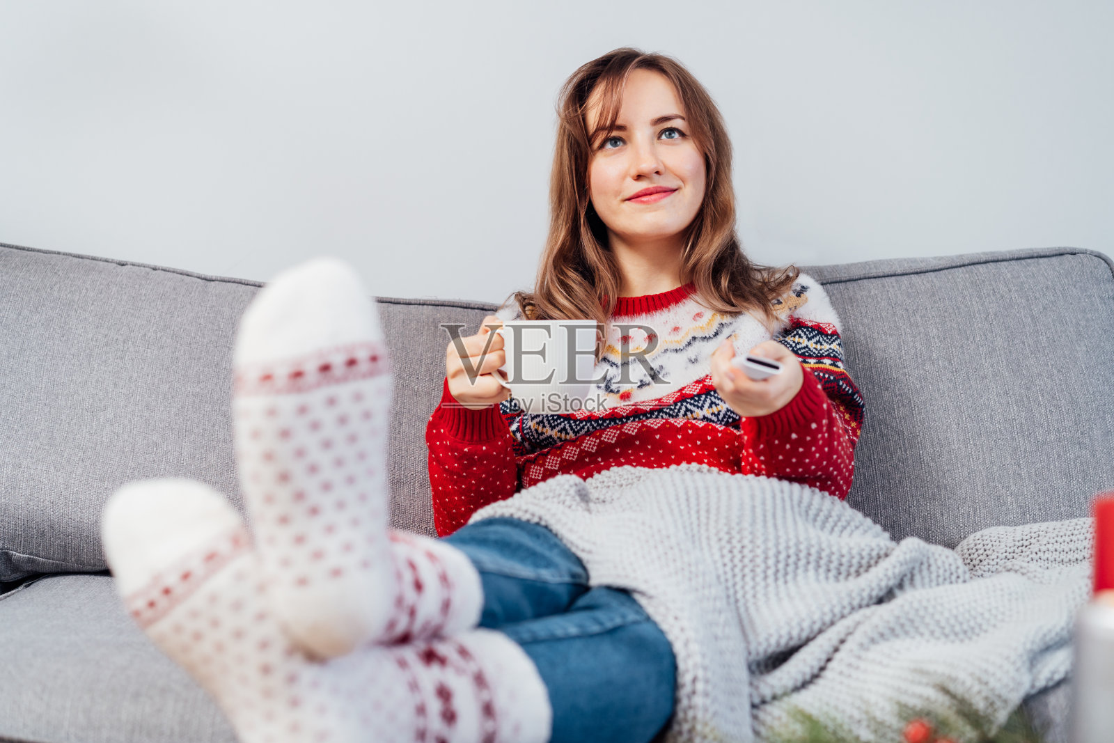 放松的女人在格纹和温暖的袜子喝热茶与遥控器在沙发上在家里看电影，电视与圣诞装饰气氛。舒适舒适的冬季概念照片摄影图片
