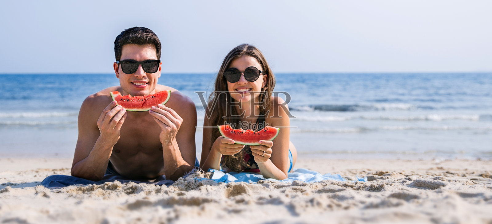 美丽的白人女子的肖像与男子在海滩上吃西瓜。新婚夫妇在海上享受蜜月。快乐的休闲情人举行在热带海滩与复制空间。照片摄影图片
