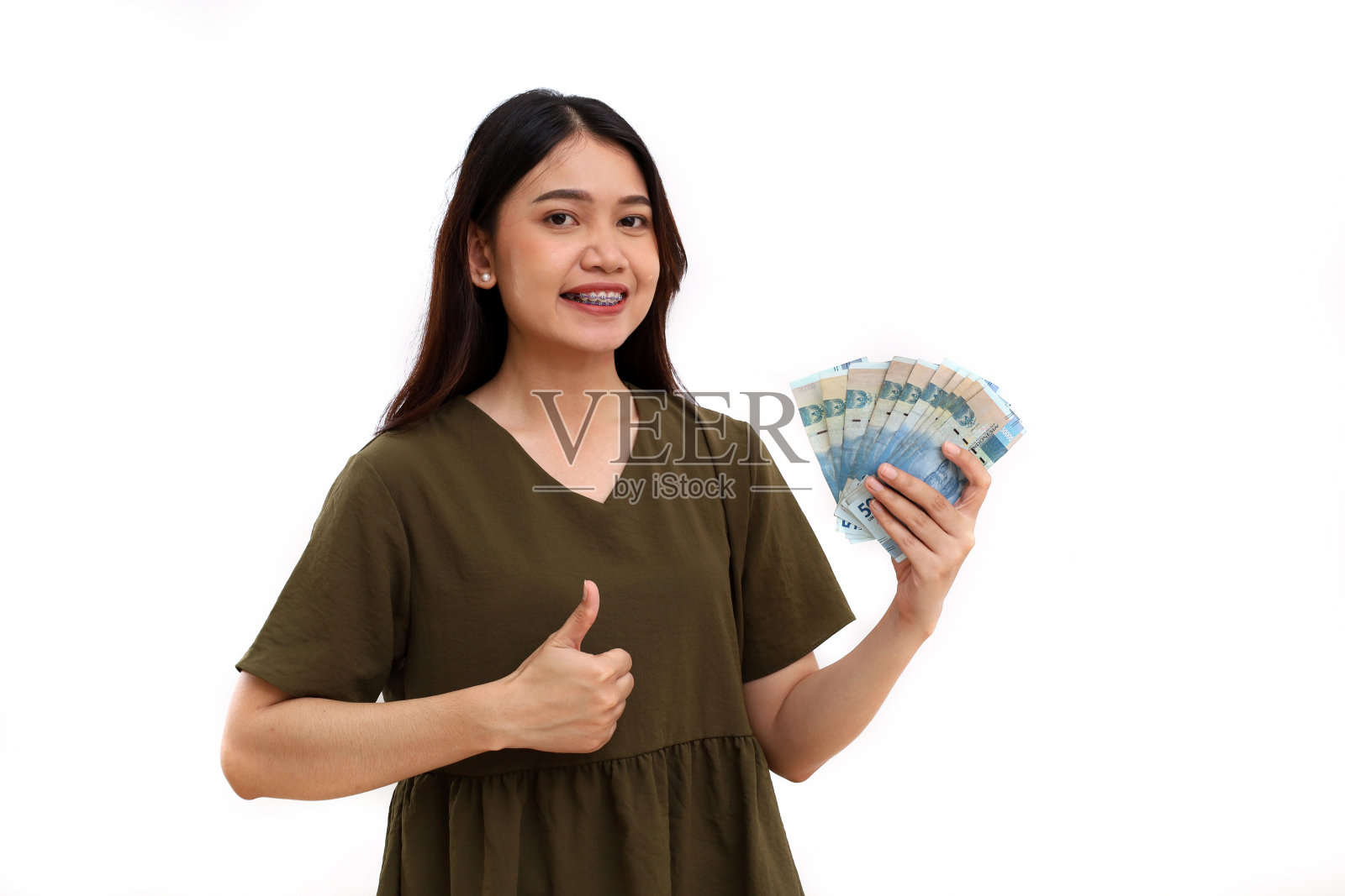 快乐的年轻美丽的亚洲女士站着，拿着印尼钞票，竖起大拇指。隔离在白色上照片摄影图片