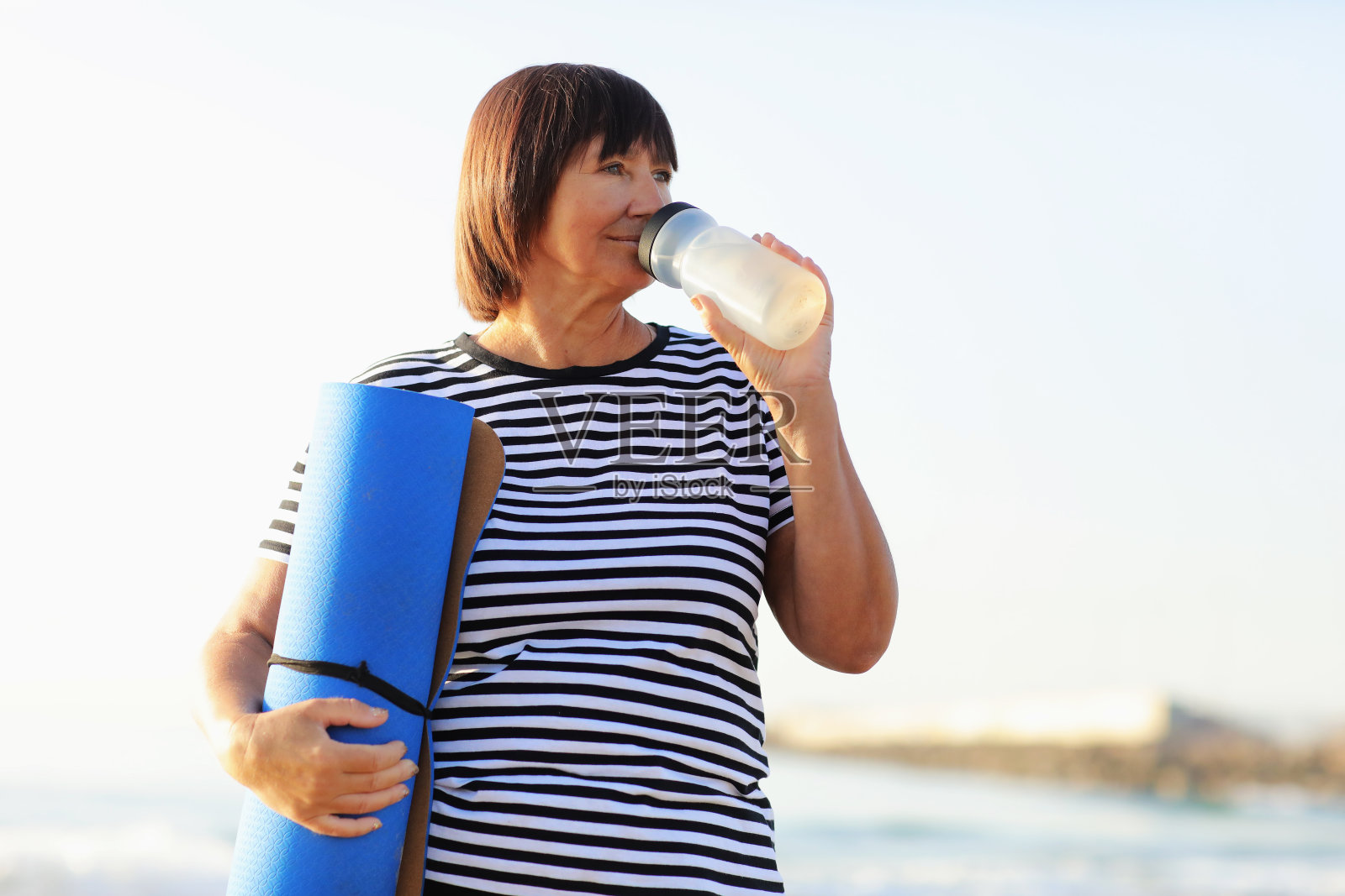 成熟女性在海边健身或瑜伽后喝瓶装水。健身和健康的生活方式概念-口渴的高级妇女与垫子保持运动后的水平衡照片摄影图片