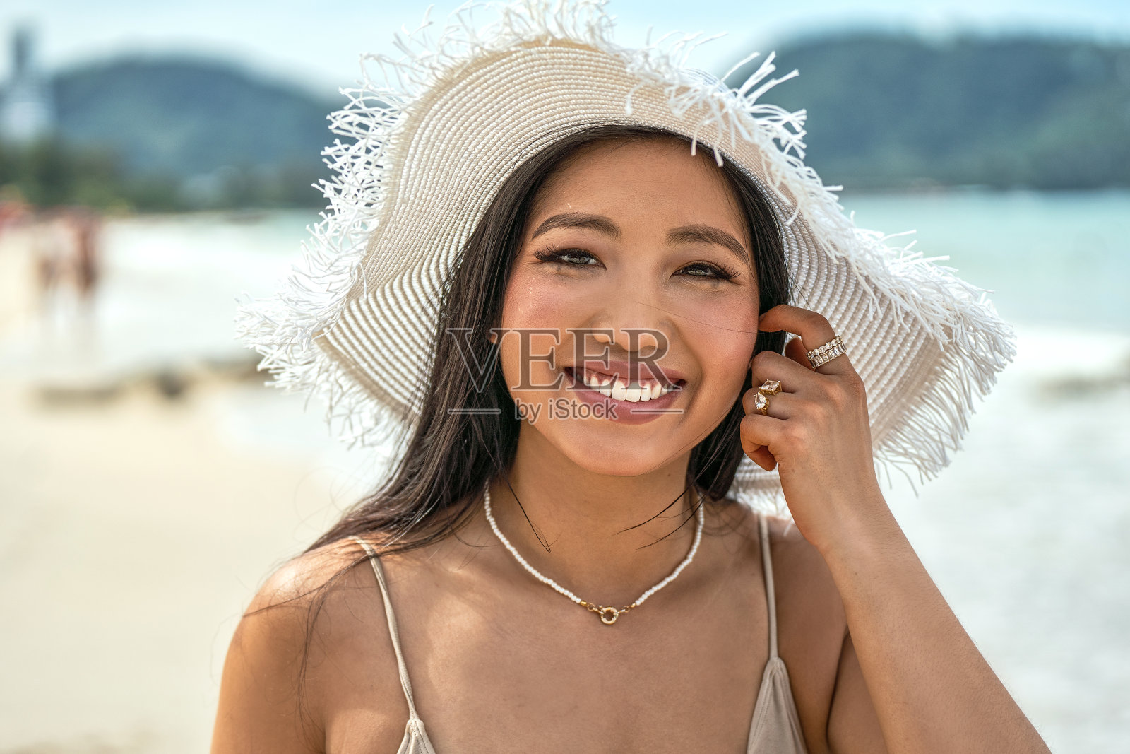 开朗美丽的女孩，带着惊人的大微笑，在热带海滩上享受阳光明媚的日子。自然女子戴着白色夏帽和首饰，看着镜头。旅游热照片摄影图片