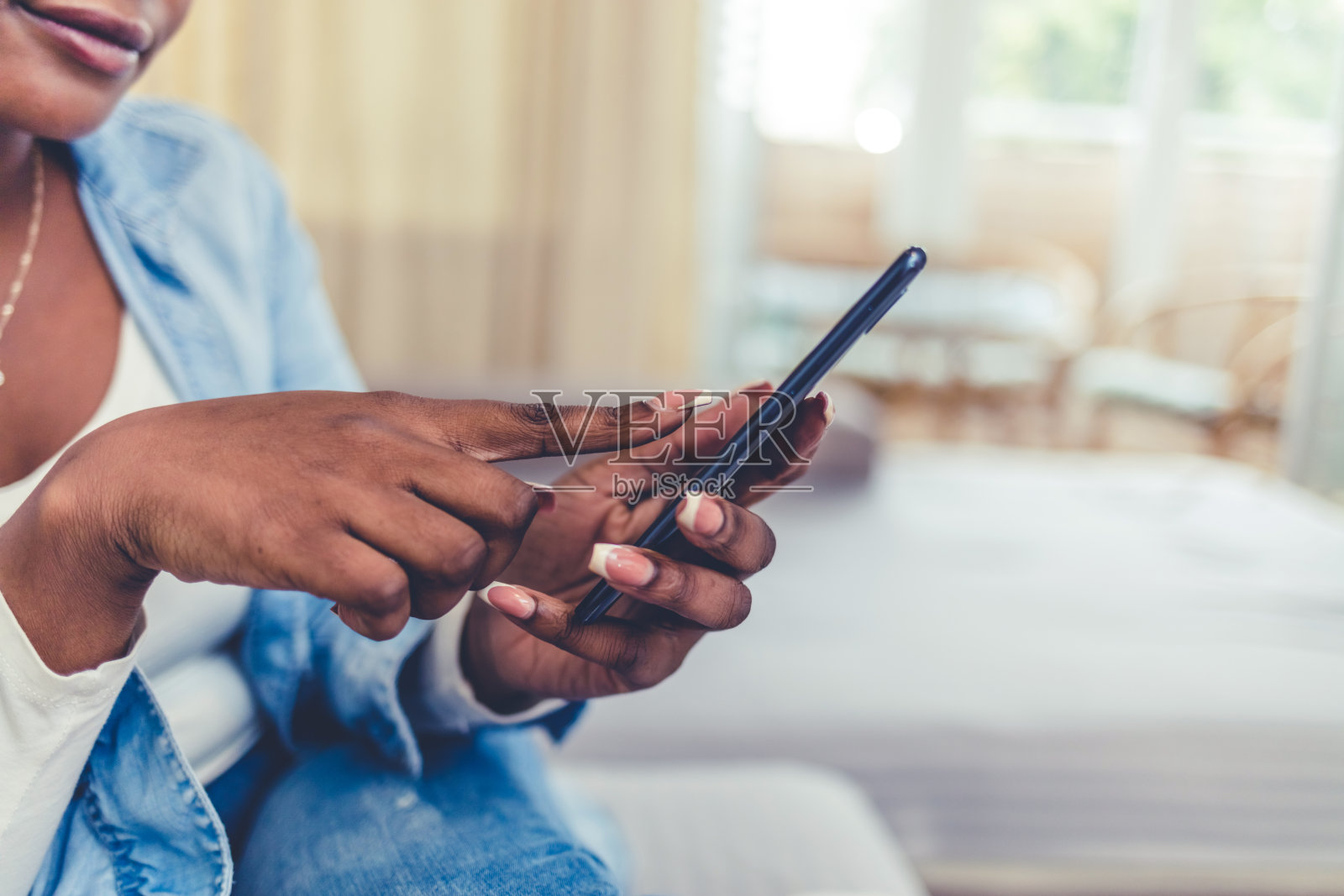 快乐的年轻非洲女子十几岁的女孩放松休息在家里的沙发上，使用智能手机，浏览新闻电子邮件，在线社交媒体，与朋友聊天。照片摄影图片