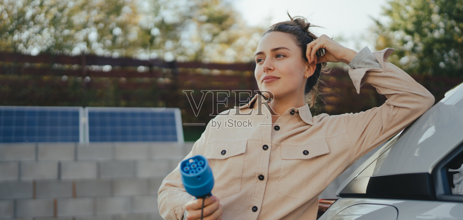年轻女子从她的电动汽车上拿着电源电缆，准备在家里充电，可持续和经济的交通理念。照片摄影图片