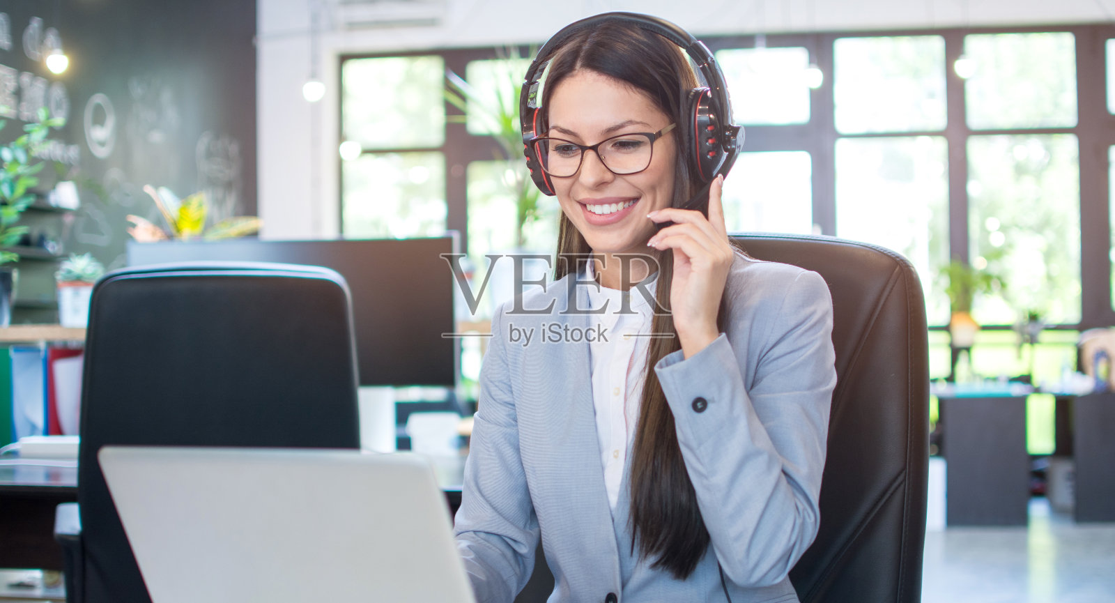 年轻的女商人戴着耳机在现代办公室的笔记本电脑上工作。照片摄影图片