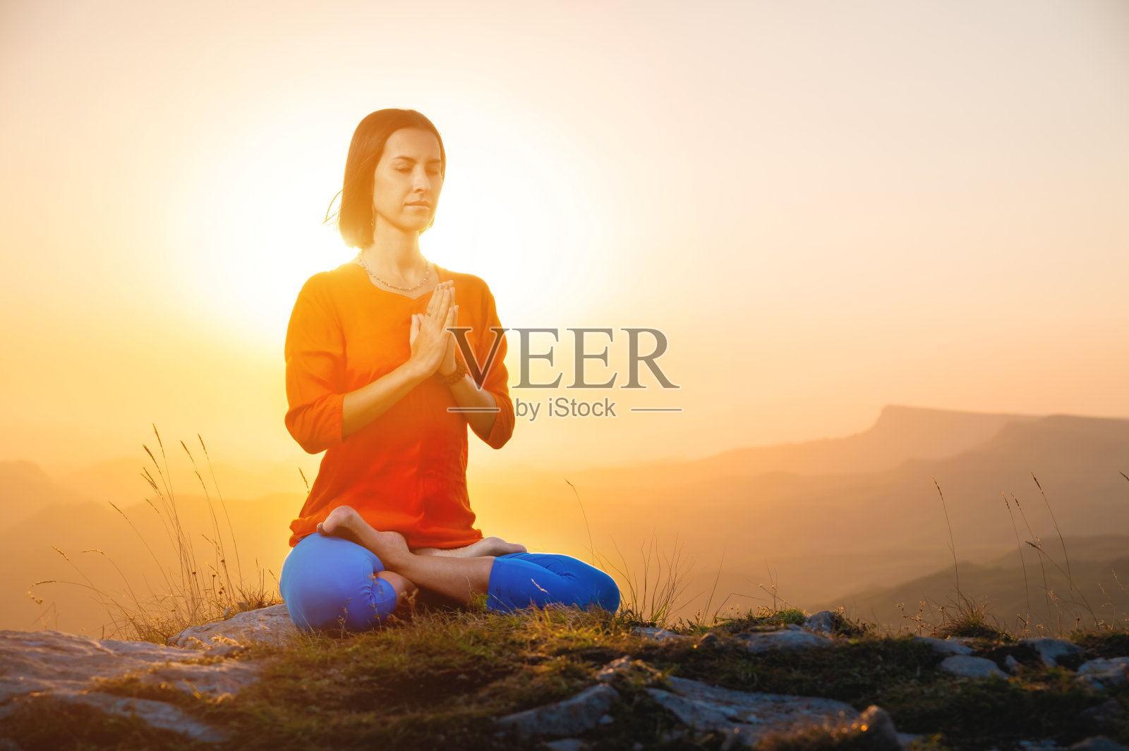 瑜伽修行者以莲花的姿势，面朝大地，坐在悬崖上，以日落的天空和群山为背景，祈祷照片摄影图片