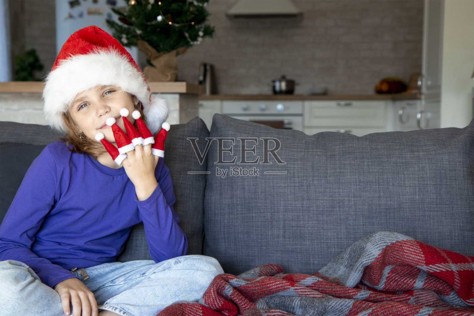 一个戴着圣诞老人帽子的金发小女孩坐在客厅的沙发上。手指上戴着圣诞老人的小帽子。圣诞和新年游戏。放置文本的地方照片摄影图片