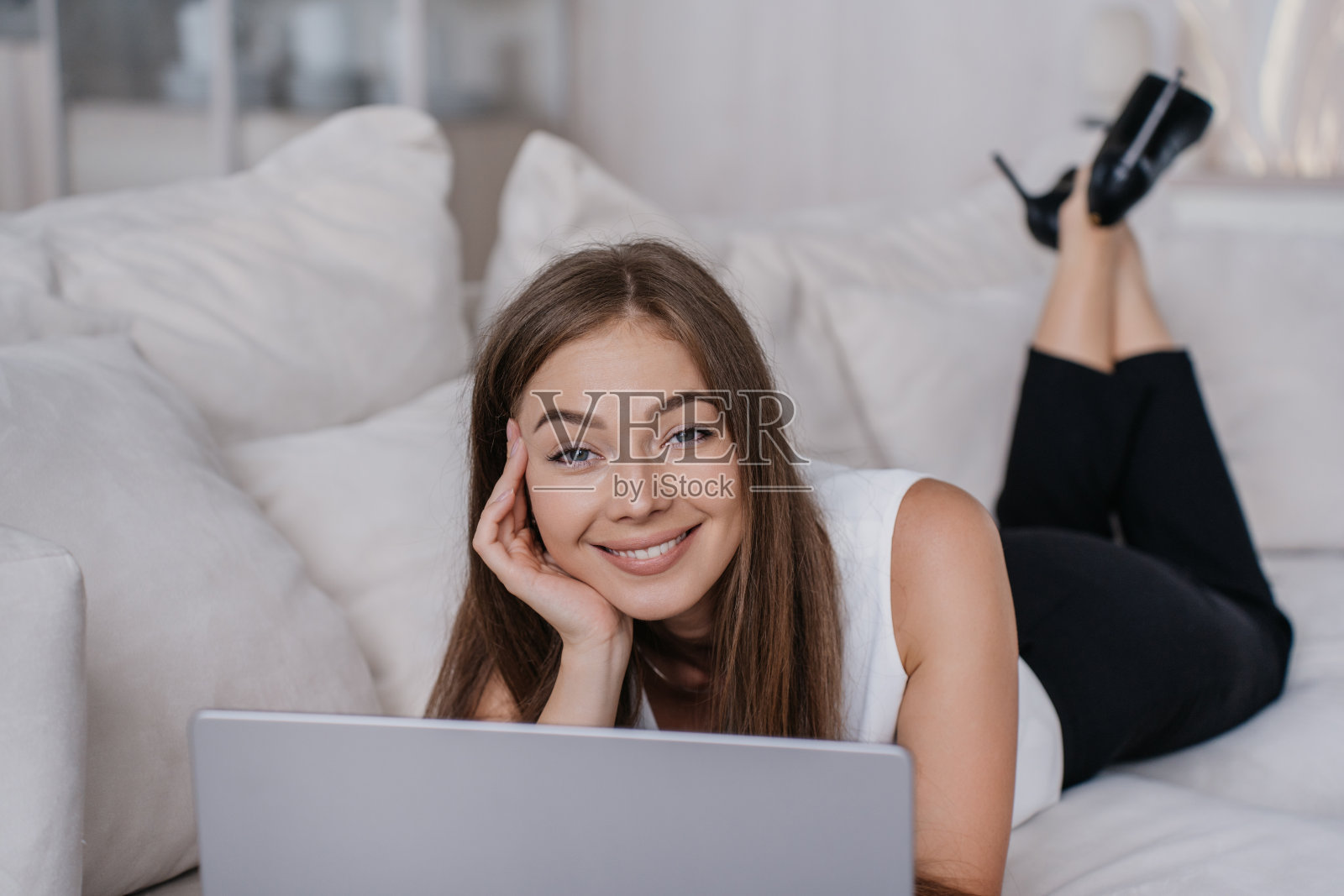 穿着白衬衫和黑裤子的白人年轻女子躺在沙发上，手里拿着笔记本电脑，对着镜头露齿微笑。华丽的意大利金发学生远程学习使用互联网。商业和教育。照片摄影图片