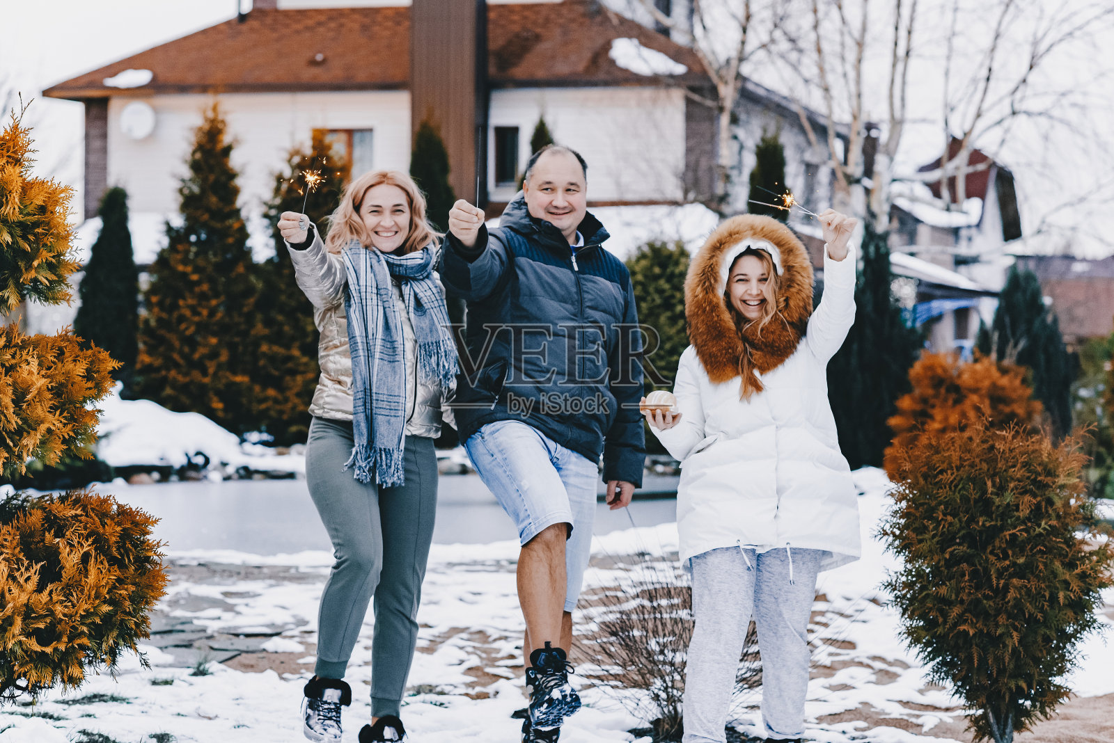 兴高采烈的爸爸妈妈和成年的女儿在冬天的大街上过新年，都站在一棵松树旁，背景是一个池塘，一片冬日的乡村风景。照片摄影图片