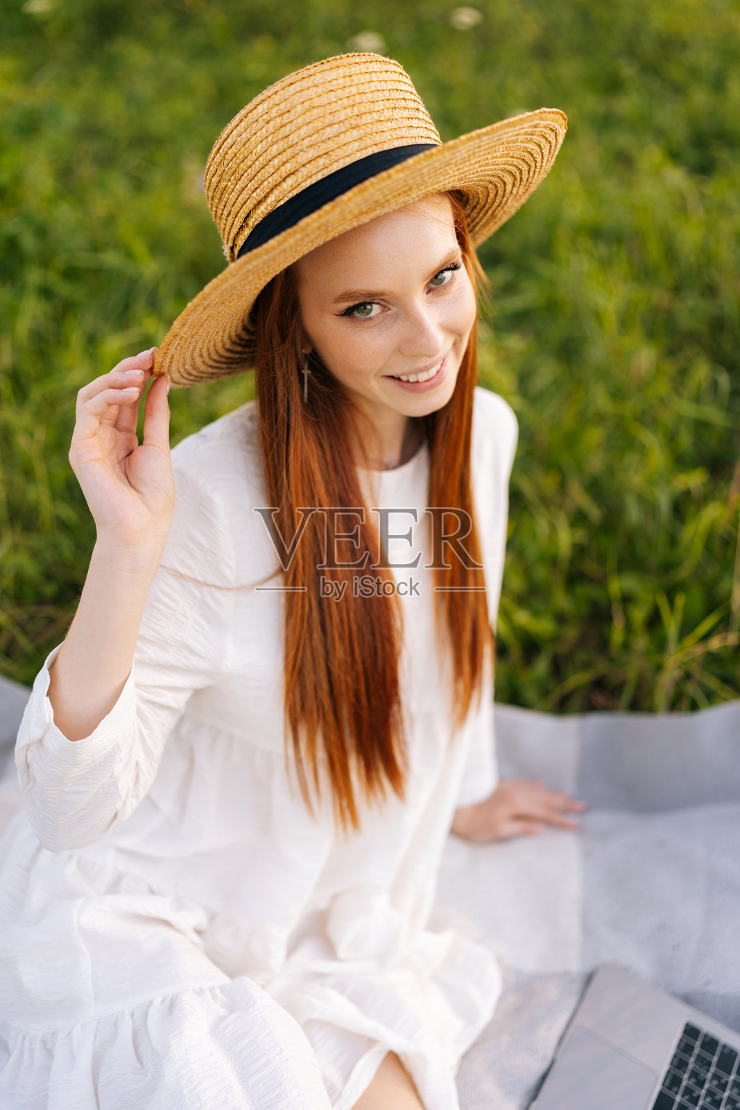 垂直裁剪拍摄漂亮的微笑年轻的姜色女子在草帽和白色连衣裙坐在摆造型的格纹在笔记本电脑看着相机。照片摄影图片