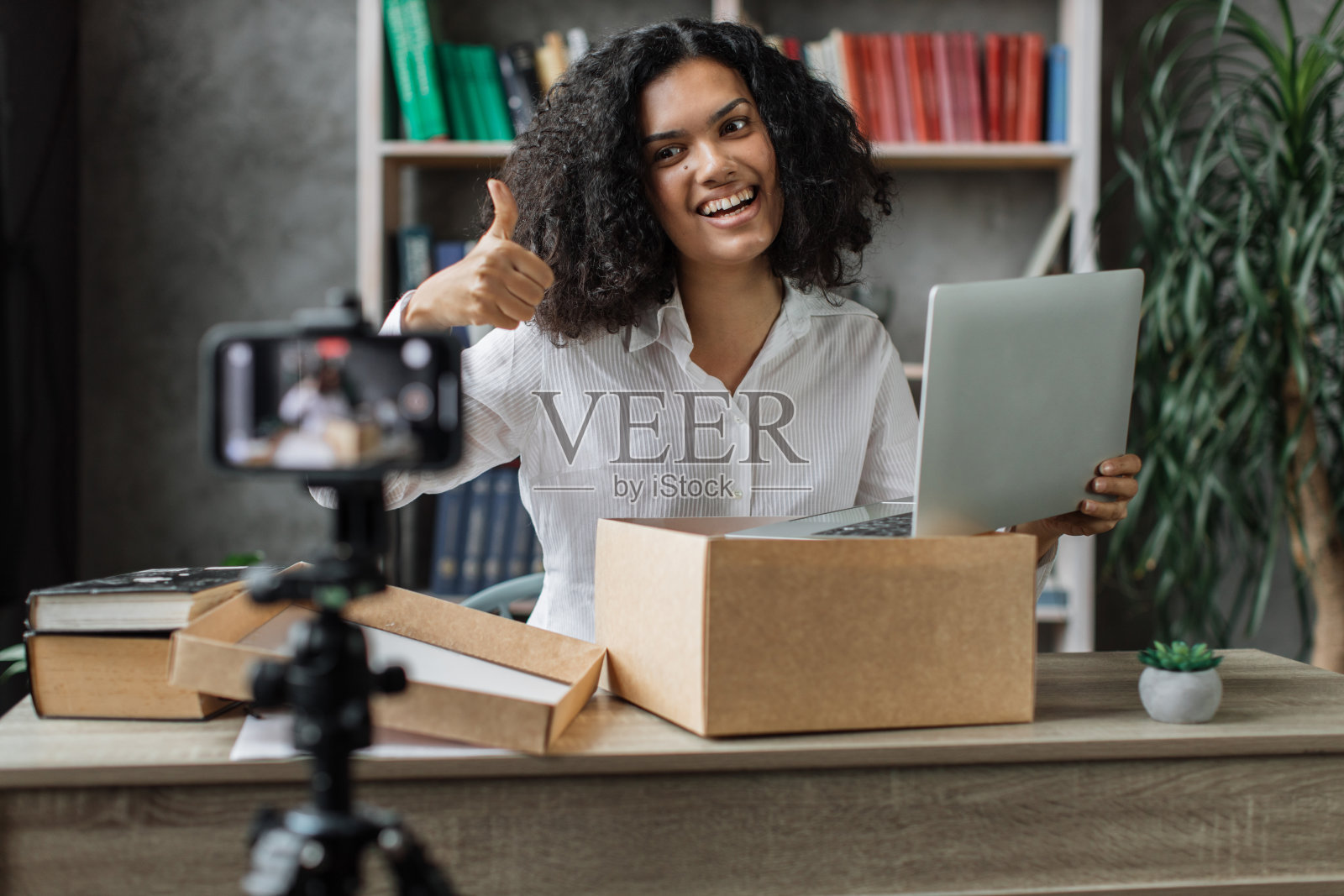 多元文化的妇女用手机摄像头录制视频，同时用新的无线笔记本电脑打开包装盒。照片摄影图片