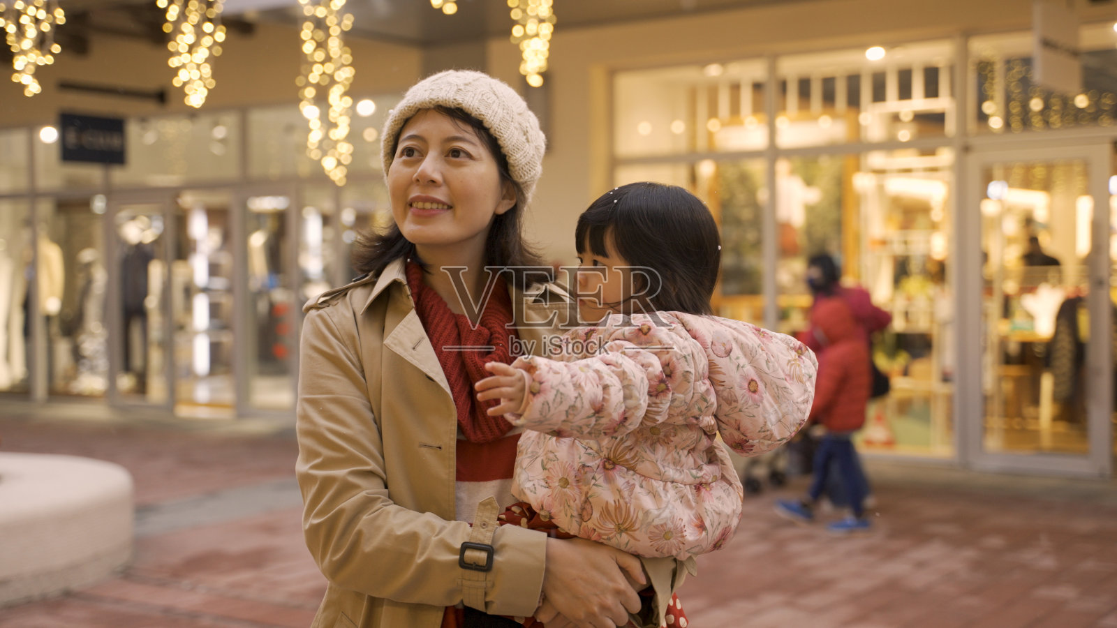 在灯火辉煌的城市街道上，快乐的亚洲妈妈举起手，看着雪花在空中飞舞，孩子抱在怀里好奇地指指点点照片摄影图片