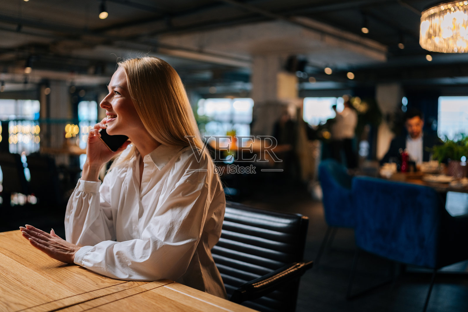 积极美丽的金发女性在时尚的衣服上与男友用智能手机聊天，坐在餐厅里，有黑暗的现代内饰。照片摄影图片