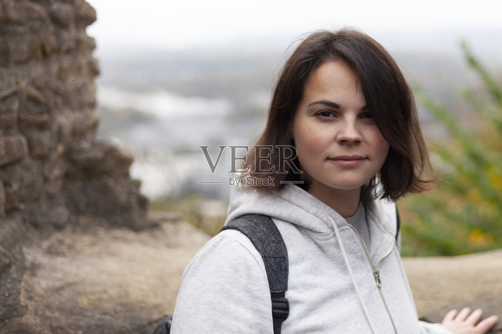 一个穿着连帽衫、背着双肩包的美丽女孩站在堡垒废墟的背景下，身后是模糊的风景。照片摄影图片