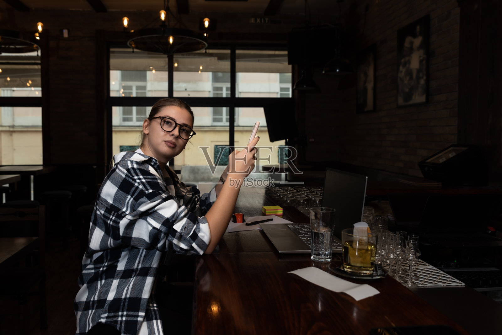 年轻的自由职业商业女性坐在咖啡厅的吧台上，在她的笔记本电脑上工作，休息一下，在她的智能手机上玩社交媒体。工作之余的专业商务人士。照片摄影图片