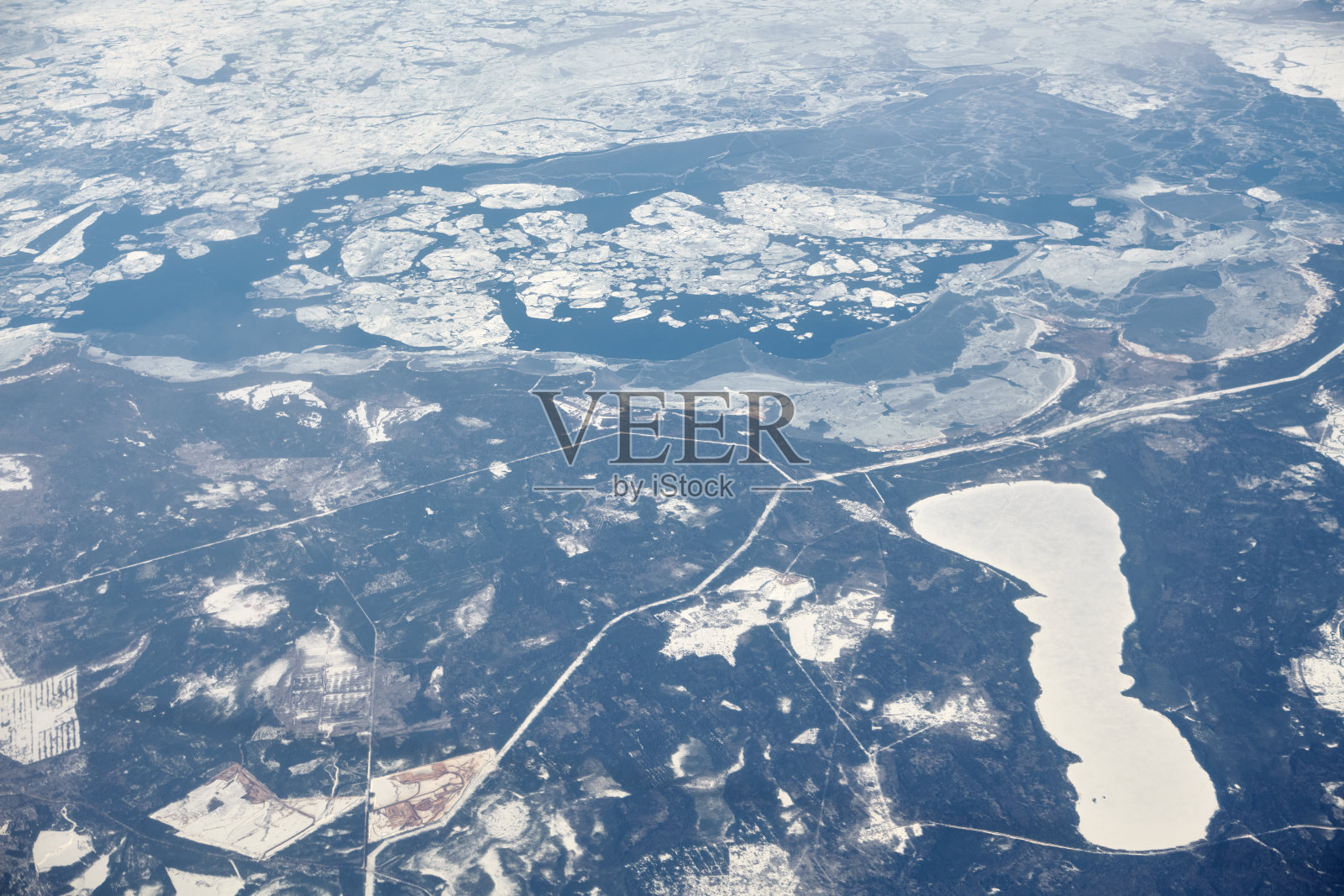 从云层上方的飞机窗口鸟瞰冰雪覆盖的河流、田野和道路照片摄影图片