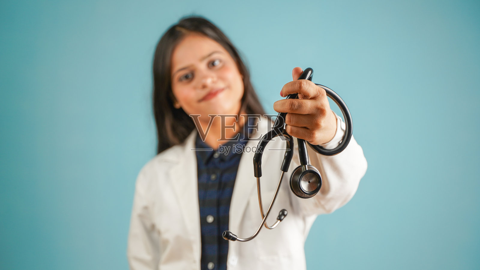 一个年轻的女医生手持听诊器的肖像，开朗的亚裔印度女医生在围裙和听诊器孤立的蓝色工作室背景照片摄影图片