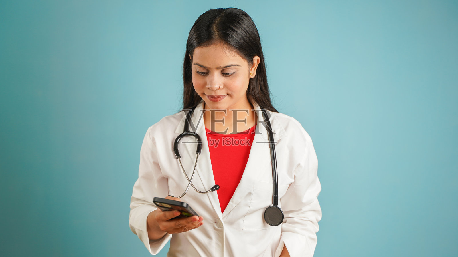 一个年轻的女医生拿着手机在她的手中，亚裔印度女医生在围裙隔离在蓝色的工作室背景，使用智能手机照片摄影图片