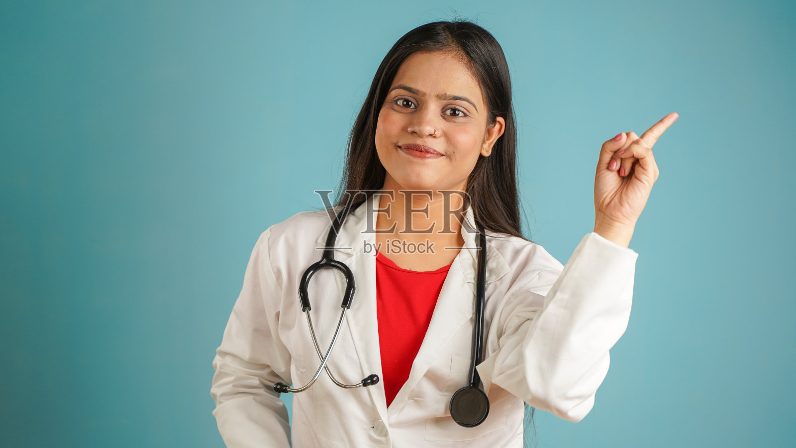 一个年轻的女医生指着手指拷贝空间的肖像，开朗的亚洲印度女医生在围裙和听诊器显示空拷贝空间孤立在蓝色工作室背景照片摄影图片