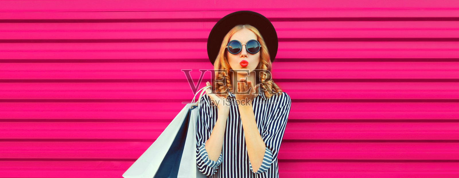 美丽的年轻女子的肖像购物袋吹她的嘴唇送飞吻穿着条纹衬衫，黑色圆帽子在粉红色的背景照片摄影图片