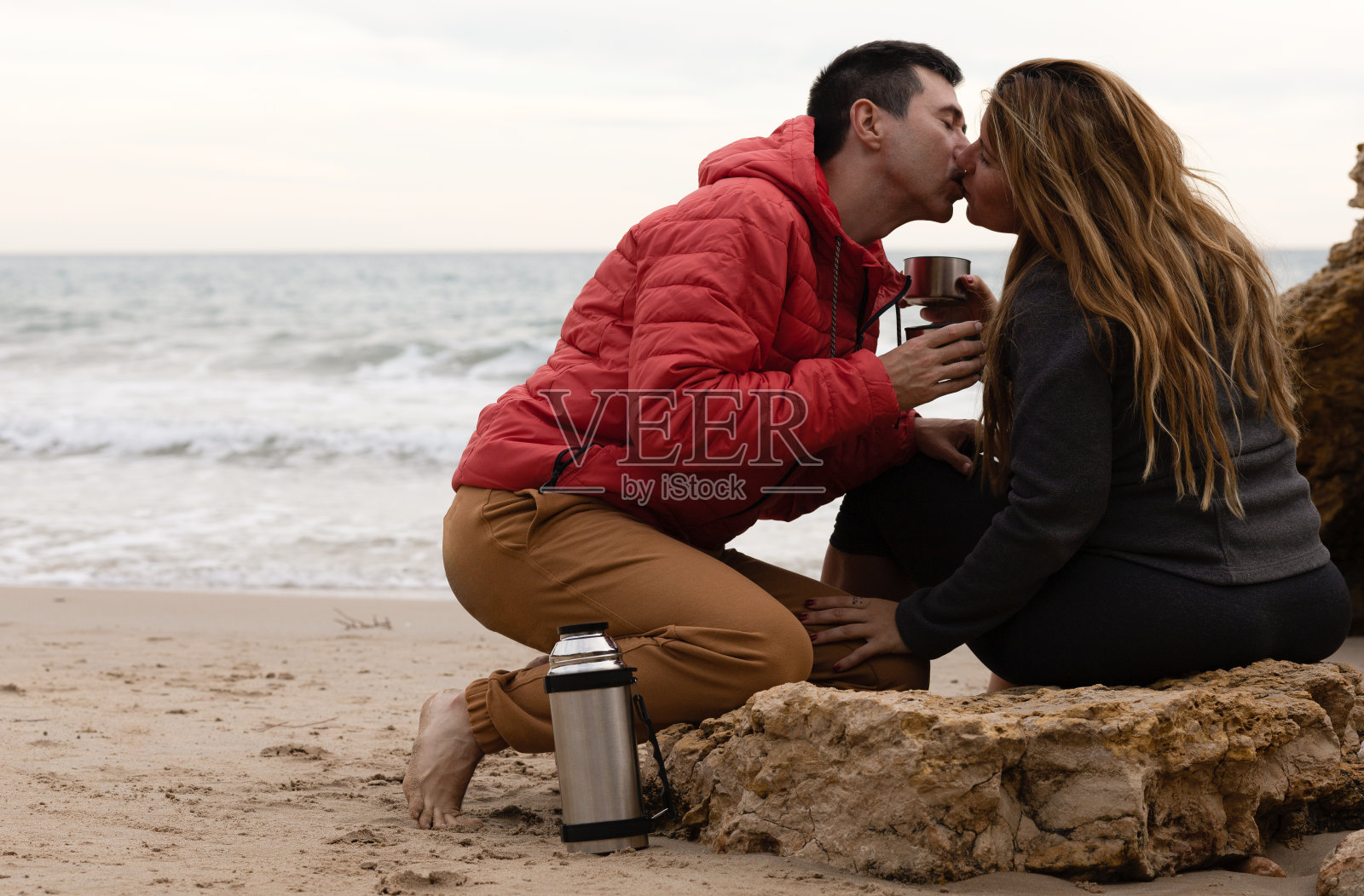 一对情侣在可重复使用的环保瓶子里接吻、喝咖啡。他们在大海面前很开心。照片摄影图片