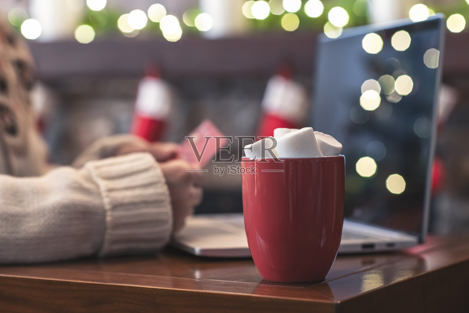 女人拿着信用卡，用笔记本电脑做订单，坐在桌子上，一杯热可可和棉花糖在圣诞壁炉与灯泡装饰。关闭了。照片摄影图片