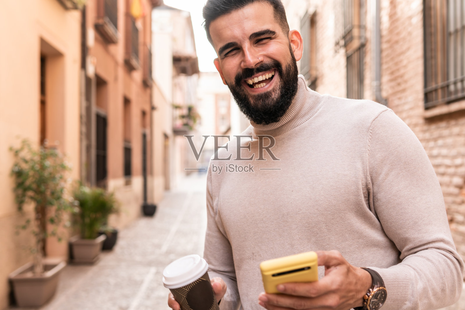 一个留着胡子、穿着高领毛衣的年轻人一边看着镜头，一边一手拿着咖啡，另一只手拿着黄色的手机。照片摄影图片