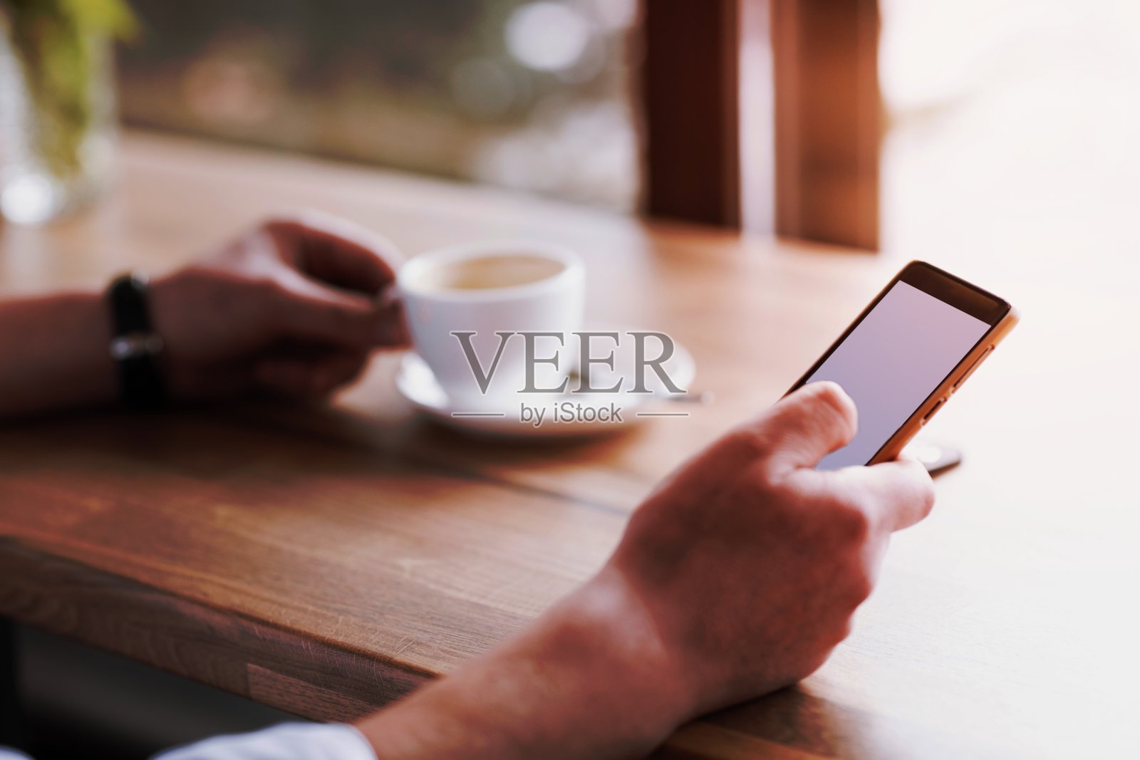 商人手里拿着手机，隔着屏幕，在咖啡店里喝咖啡，模糊的咖啡杯背景，模拟概念照片摄影图片