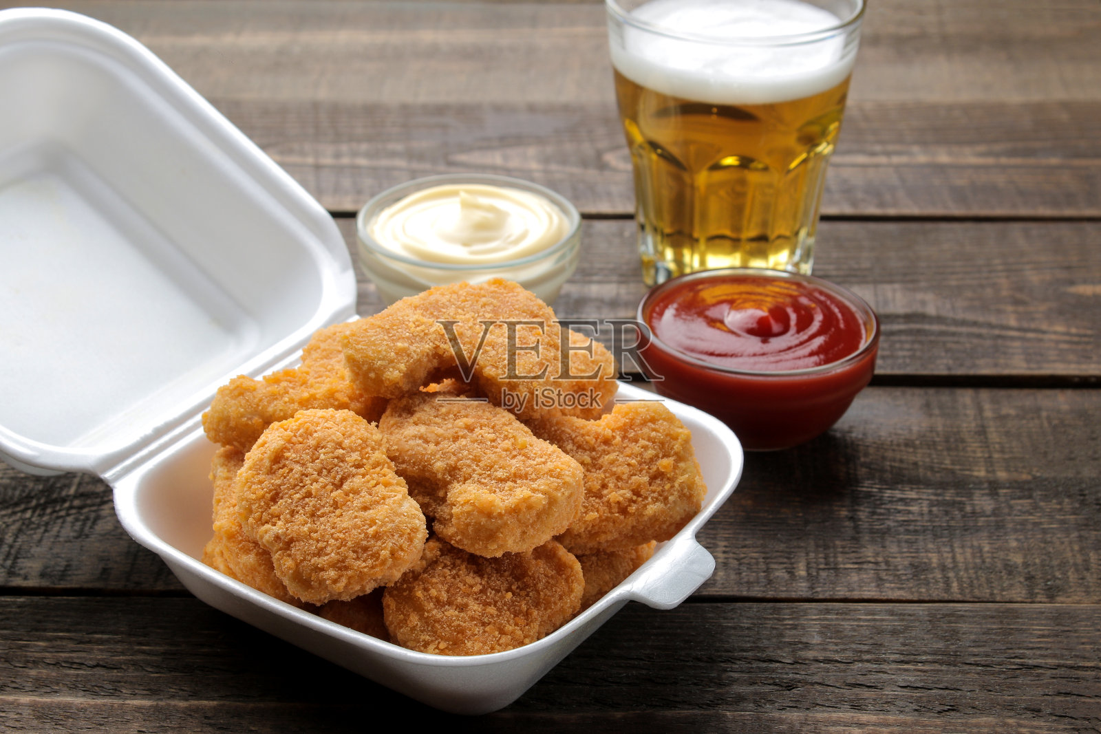 送餐盒里的鸡块，白色和红色的酱汁，棕色的木制背景上有啤酒。快餐照片摄影图片