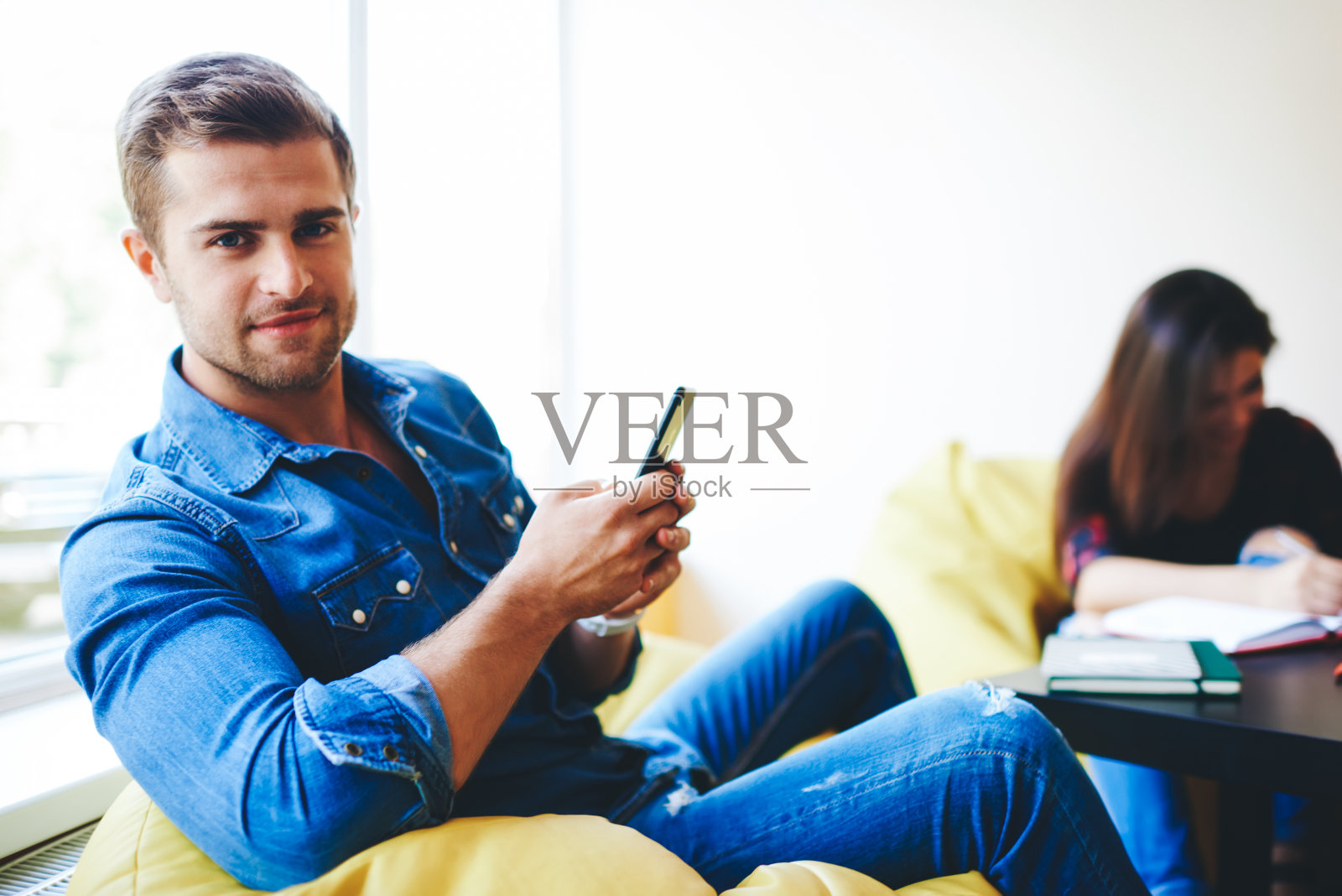 英俊的白人男性组长的肖像，使用智能手机聊天时，小组在后台合作，繁荣的男子看着相机浏览手机与朋友学习照片摄影图片