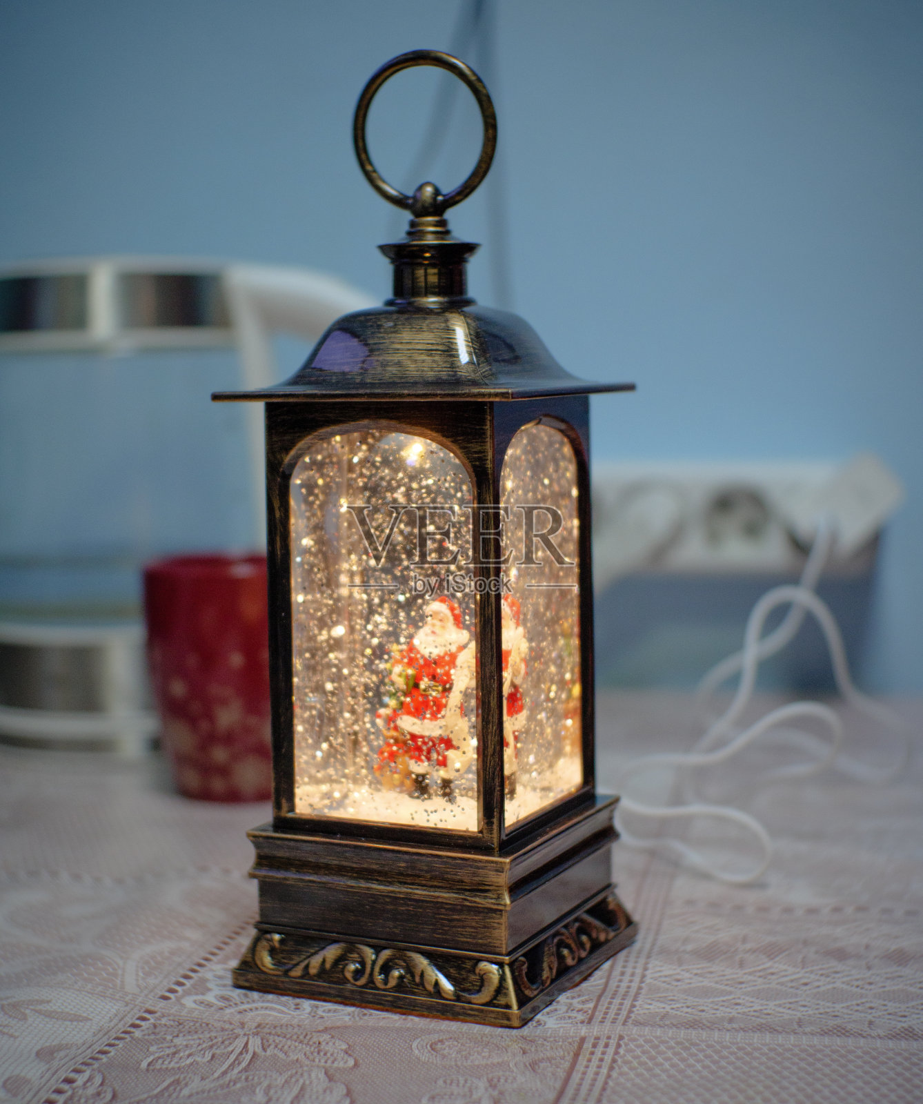 一个漂亮的圣诞灯笼在桌子上。圣诞老人站在灯笼里，雪花落在他身上。圣诞彩灯闪闪发光，营造出节日气氛。照片摄影图片