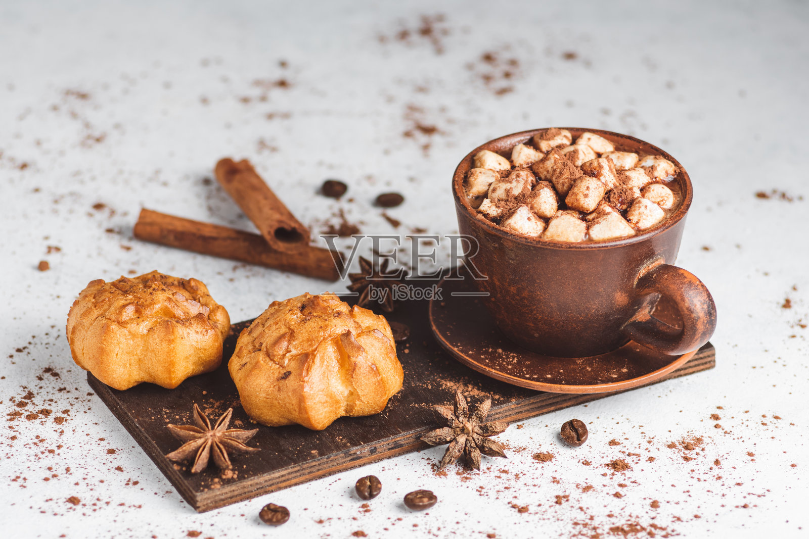 热巧克力饮料，杯子里有棉花糖，白色的桌子上有甜甜的面包泡芙，上面撒着可可粉、肉桂棒和八角茴香照片摄影图片