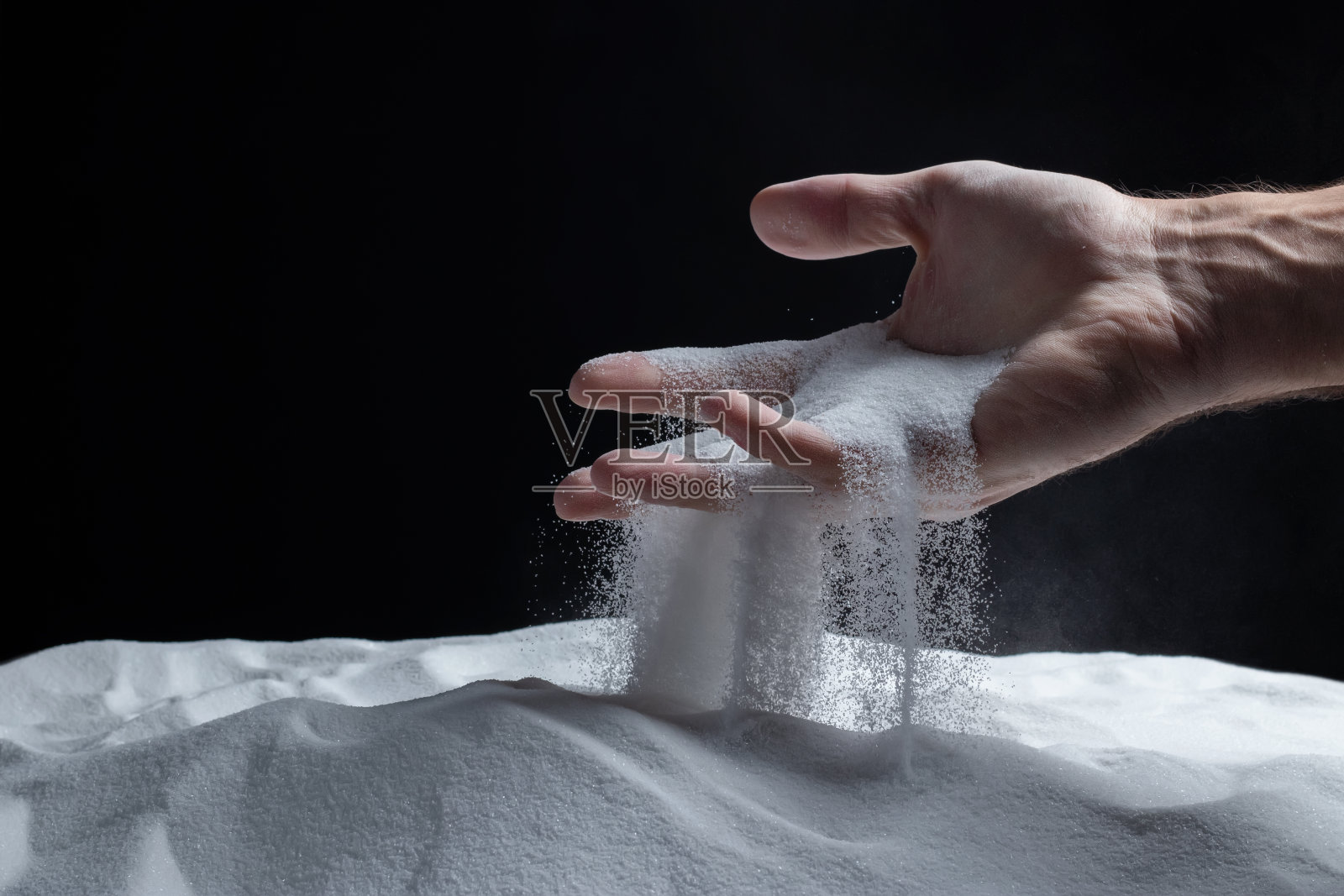 男子手里拿着一把白色的干沙子，在黑色背景上，沙子从手指间溢出。照片摄影图片