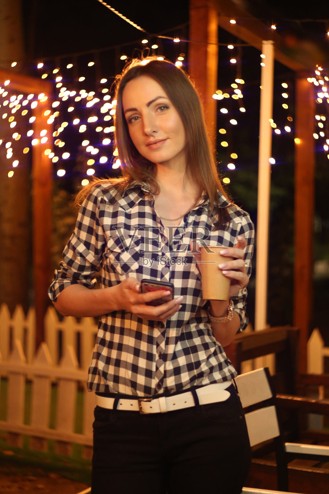 穿着格子衬衫的漂亮时髦女孩在灯笼的背景下喝着咖啡，看着手机照片摄影图片