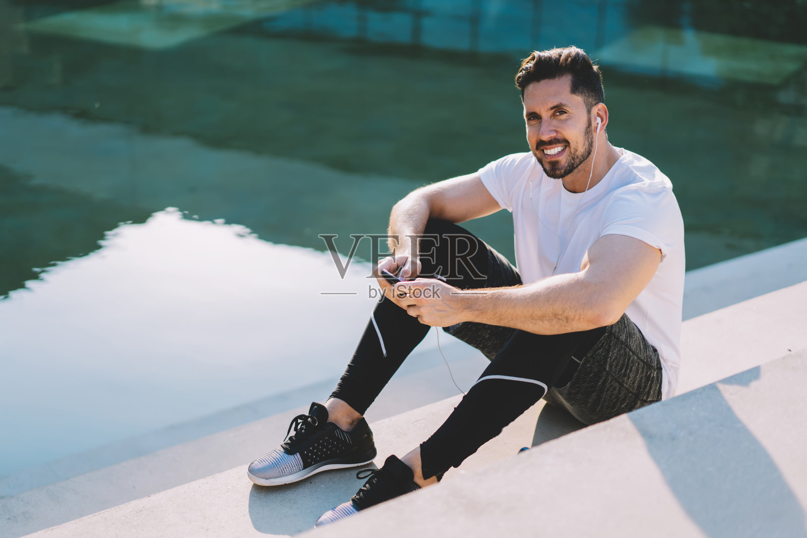 微笑的年轻运动员穿着运动服听音乐，坐在体育场上使用智能手机，20多岁的男性肌肉潮人在锻炼后休息，使用现代设备和4G连接照片摄影图片