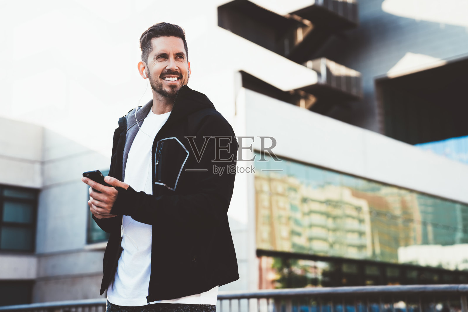 快乐的男运动员穿着时髦的夹克，满足于户外锻炼，用耳机听音乐，快乐的运动员站在城市环境检查智能手机通知照片摄影图片