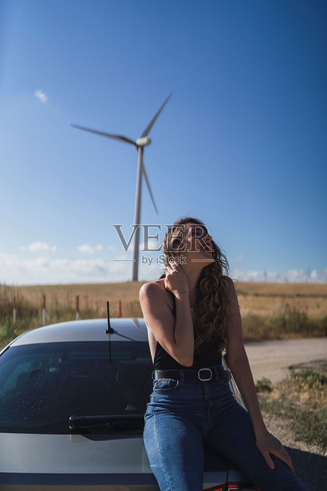 一个美丽的白人女性靠在一辆有风力涡轮机的汽车上的垂直镜头照片摄影图片