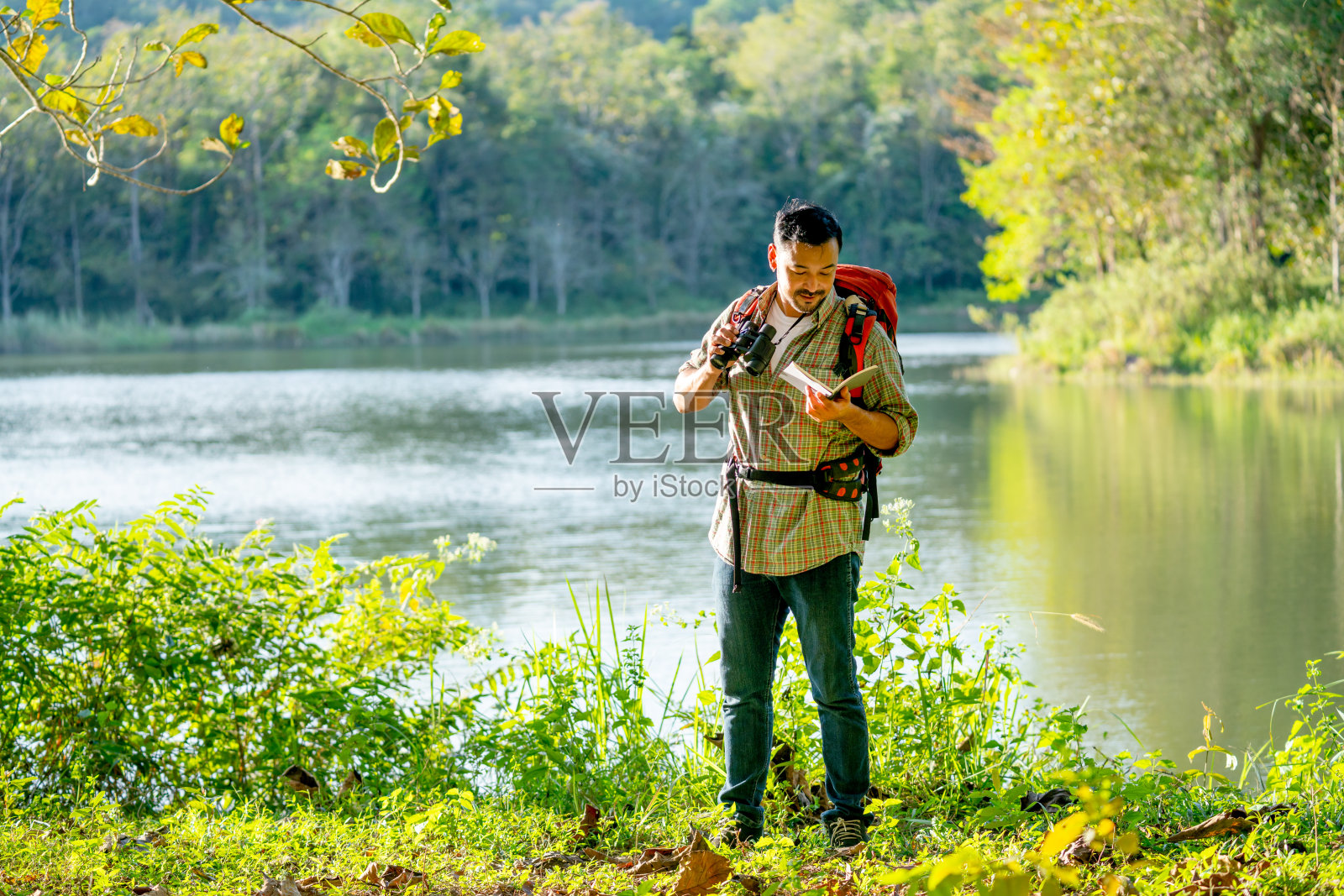 一名亚洲男子站在那里看记录了一些数据的书，他还拿着双筒望远镜，看起来他在森林里发现了一些有趣的生物。照片摄影图片