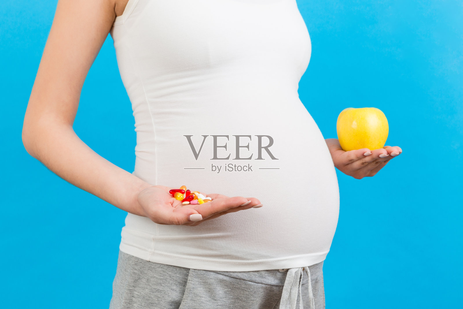 特写孕妇在苹果和一堆药丸之间做出选择，彩色背景与复制空间。怀孕期间服用维生素的概念照片摄影图片