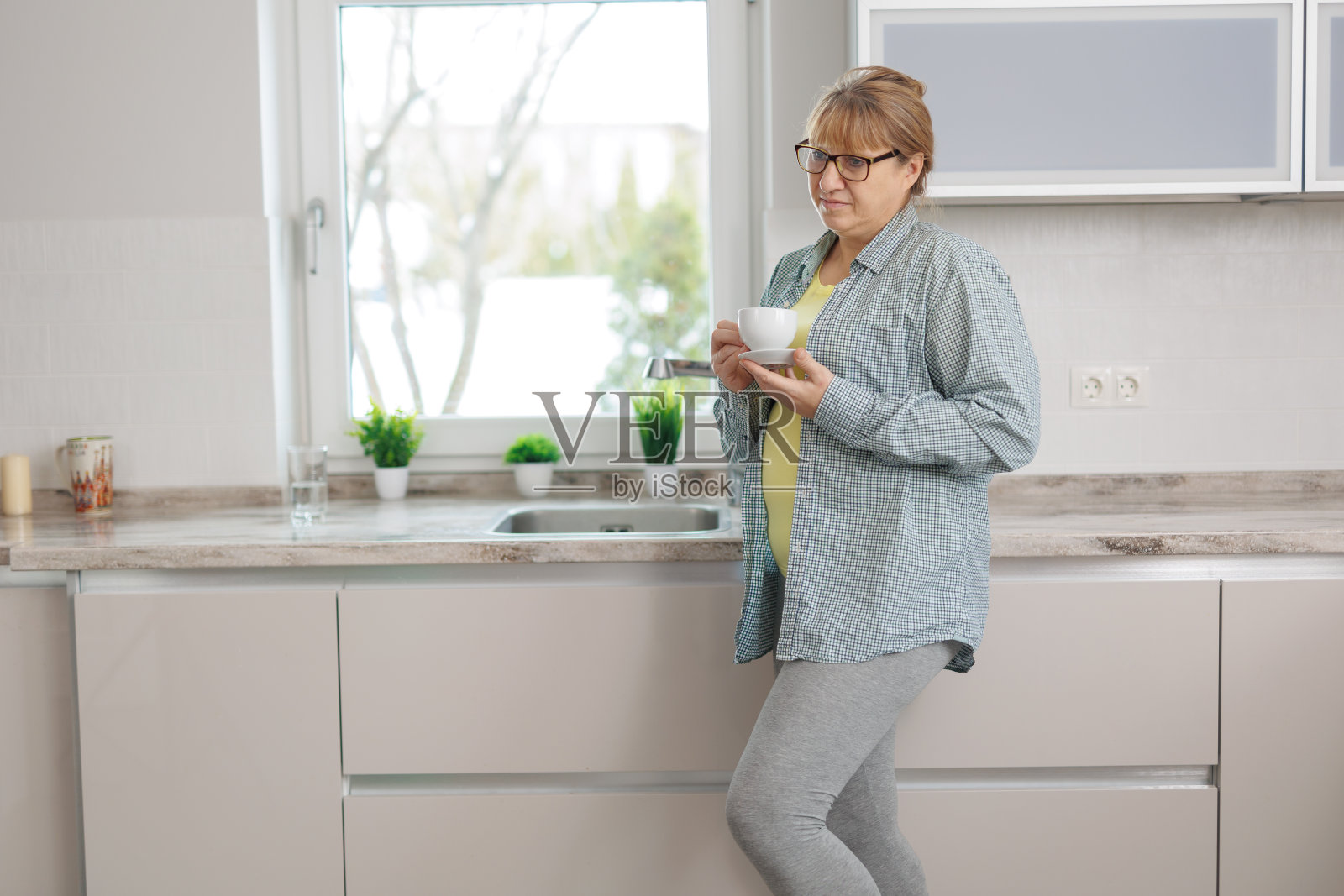 咖啡休息。冷静的高级女士在蓝色衬衫站在厨房，喝咖啡，看着空旷的空间照片摄影图片