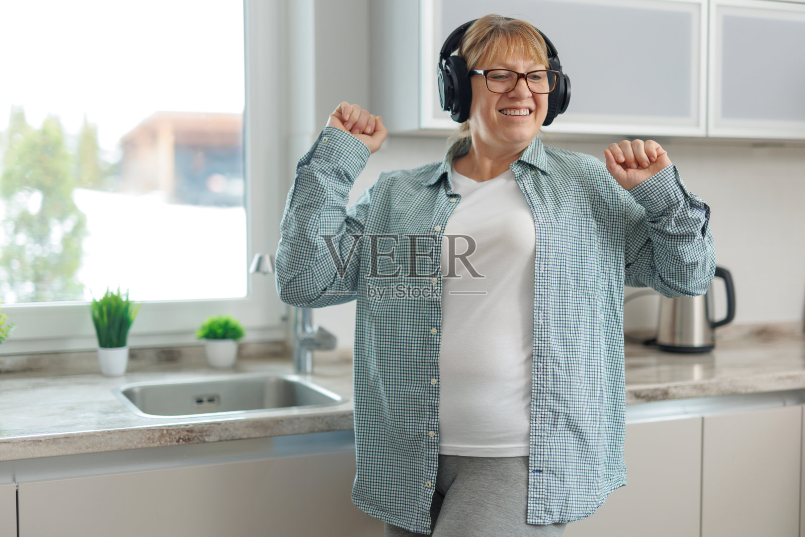 一名成年女子带着无线耳机在厨房里听音乐跳舞。一个穿着蓝色衬衫的女人微笑着跳舞。照片摄影图片