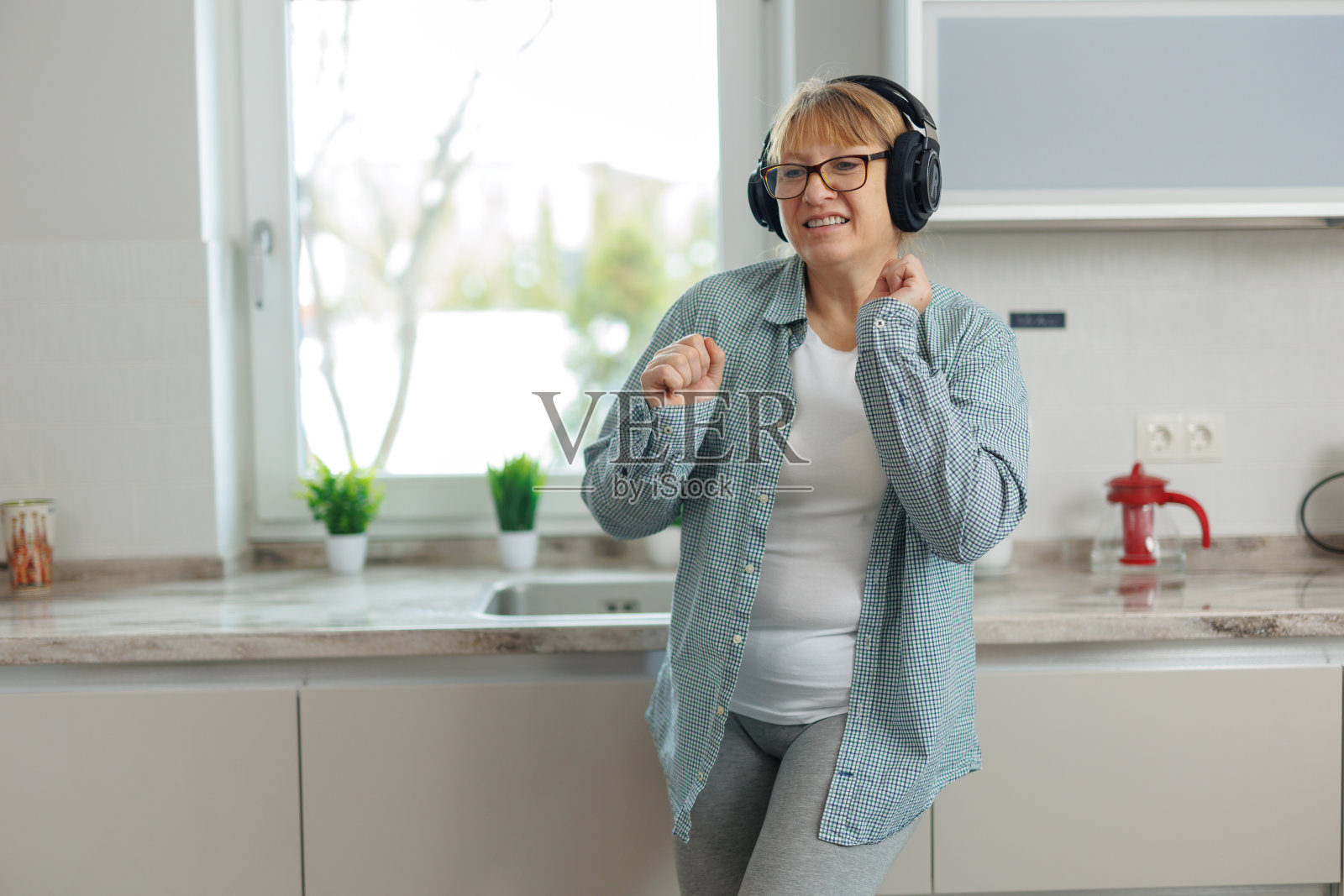 一名成年女子带着无线耳机在厨房里听音乐跳舞。一个穿着蓝色衬衫的女人微笑着跳舞。照片摄影图片