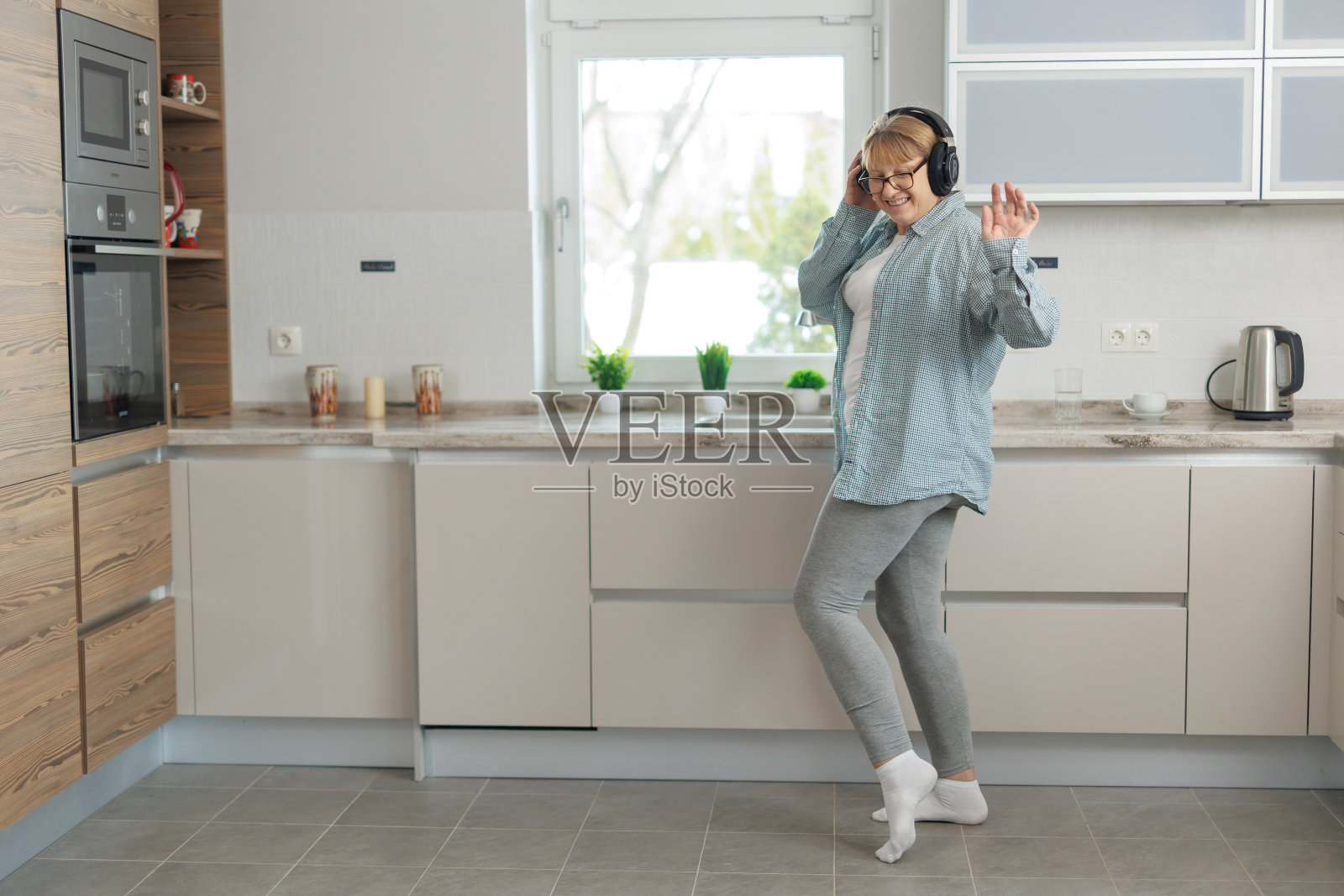 一名成年女子带着无线耳机在厨房里听音乐跳舞。一个穿着蓝色衬衫的女人微笑着跳舞。照片摄影图片